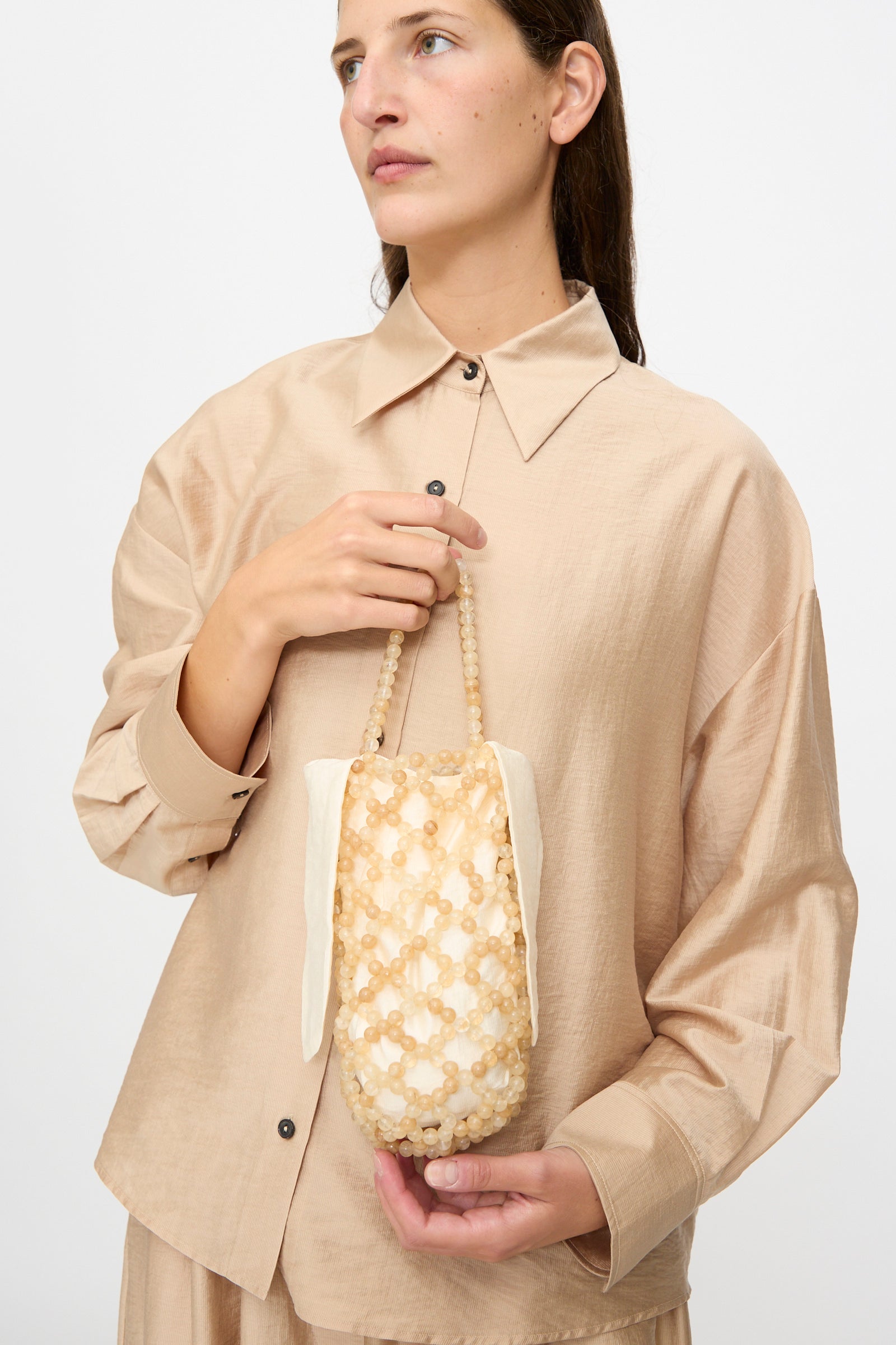 A woman in a beige shirt holds the Cordera Handmade Beaded Bag in Jasper Miel, featuring beaded details and a short handle, crafted in Guatemala, against a plain background.