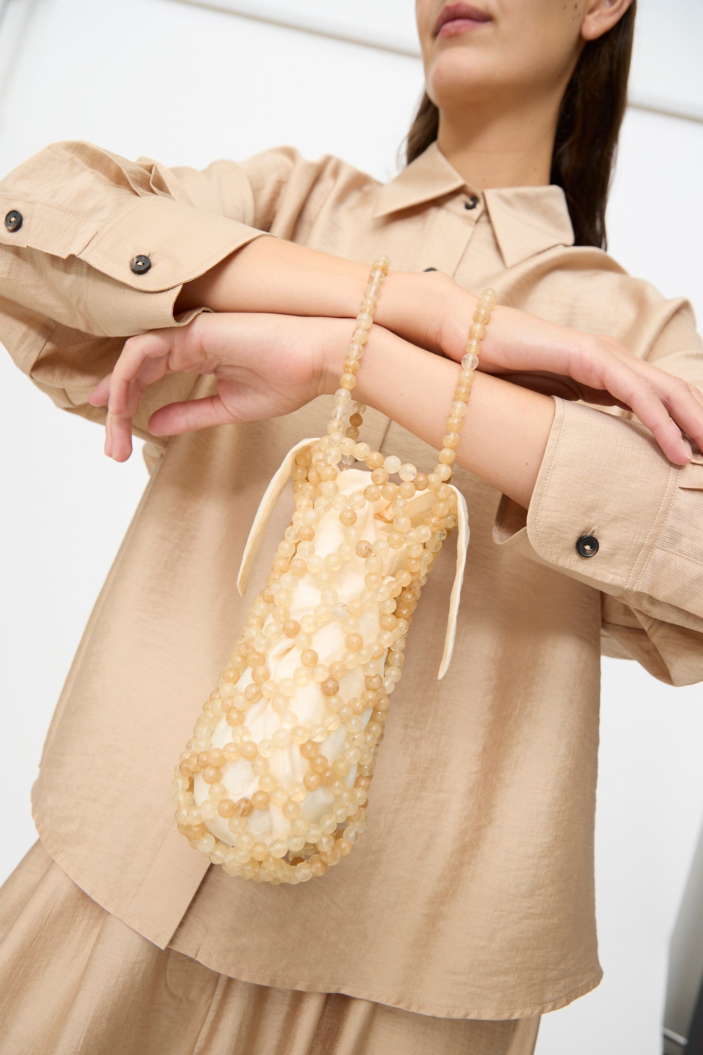 A woman in a beige outfit holds the Cordera Handmade Beaded Bag in Jasper Miel, a small, cylindrical accessory crafted from translucent beads with two handles.
