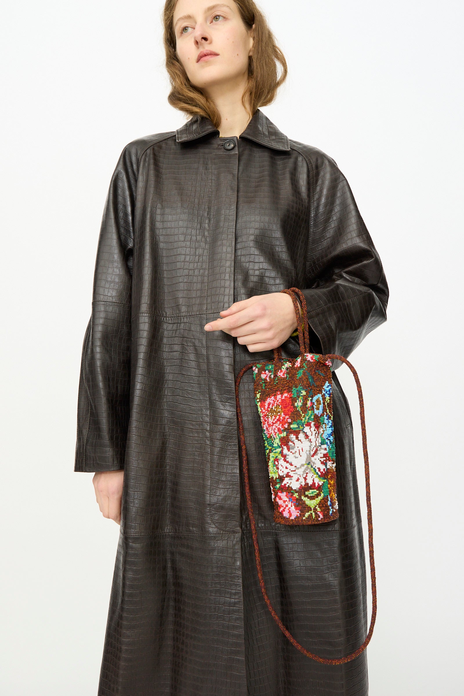 A person in a long, textured dark brown coat holds Cordera's Handmade Beaded Bag in Peonia—featuring a colorful floral design and long strap—against a plain white background.