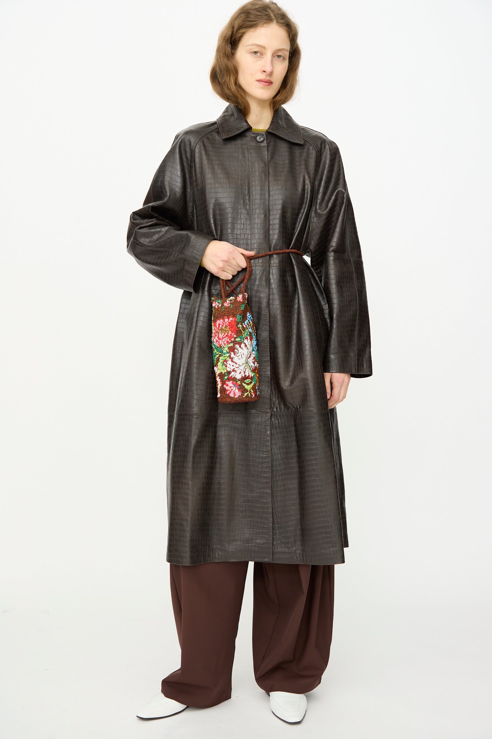A person with a long, dark textured coat, wide brown trousers, white shoes, and holding Cordera's Handmade Beaded Bag in Peonia stands against a plain background.