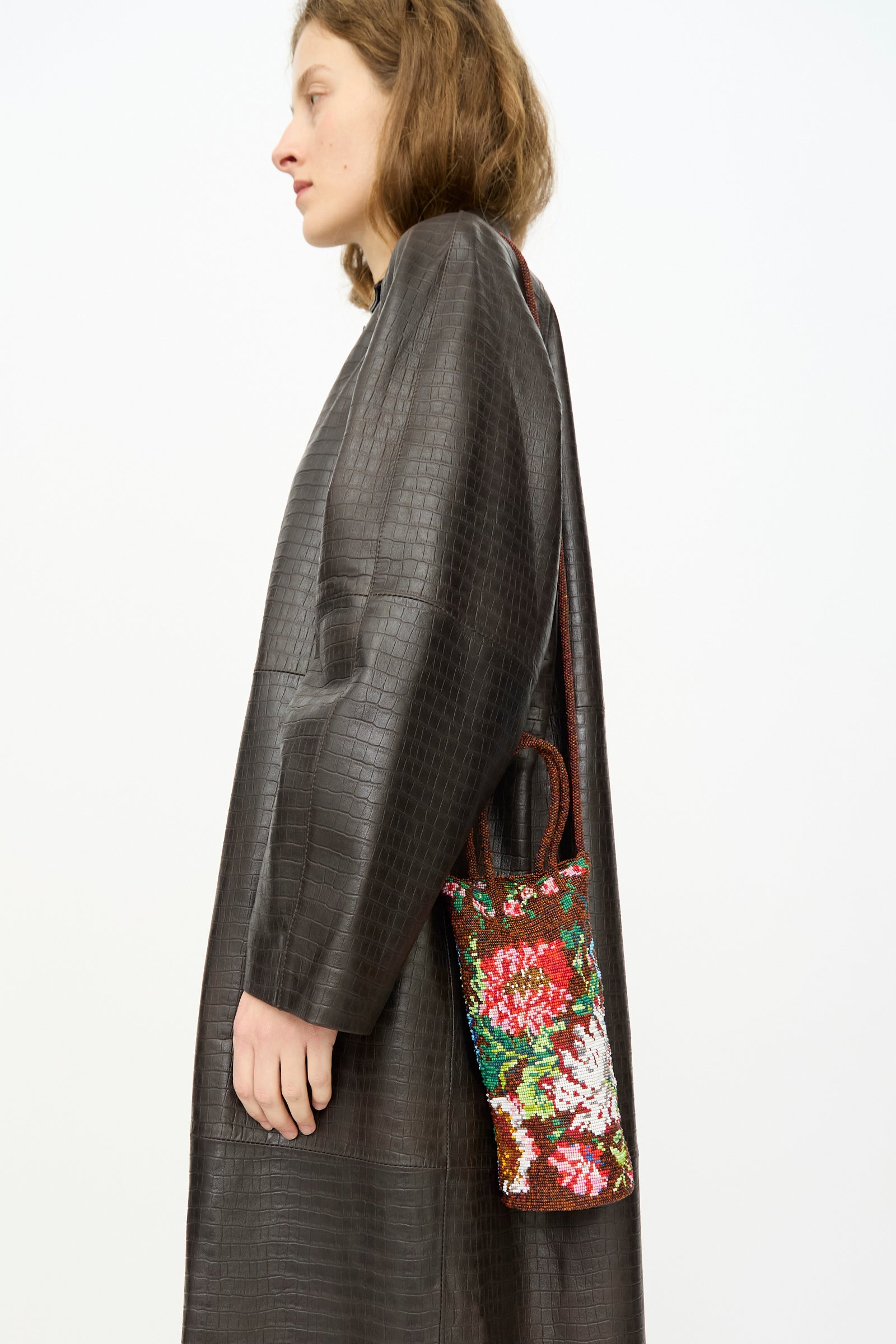 Someone in a textured dark coat holds the Cordera Handmade Beaded Bag in Peonia, featuring a shoulder strap, handcrafted in Guatemala, against a plain light background.