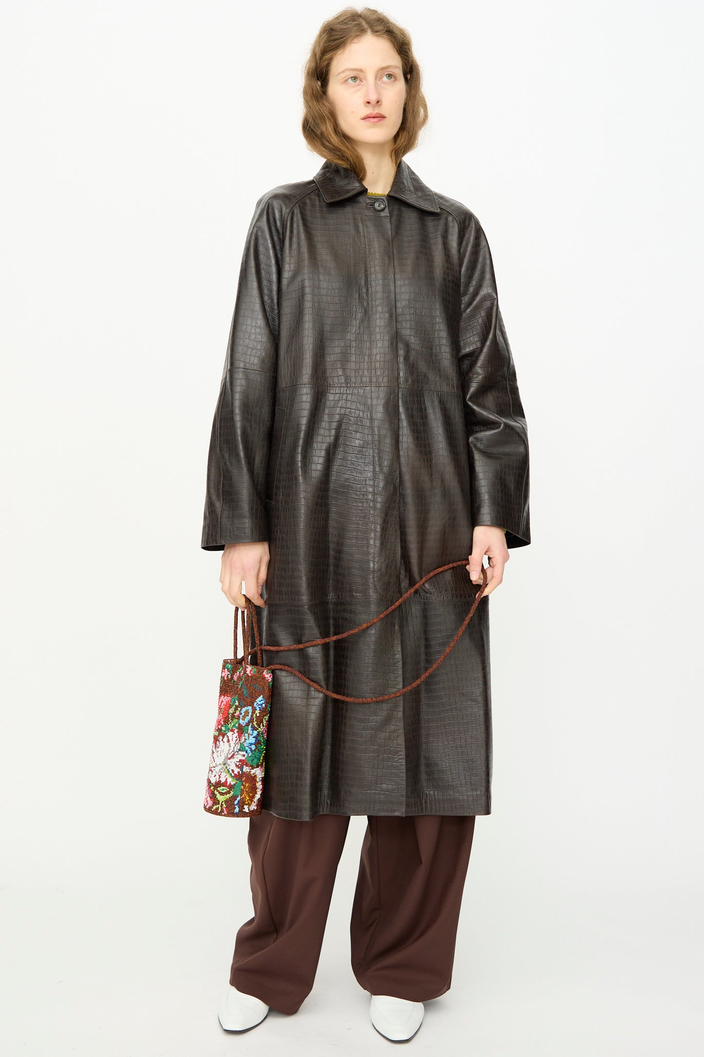 Wearing a long, dark textured coat over brown wide-leg pants, a person holds Cordera’s Handmade Beaded Bag in Peonia with white shoes, standing against a plain white background.