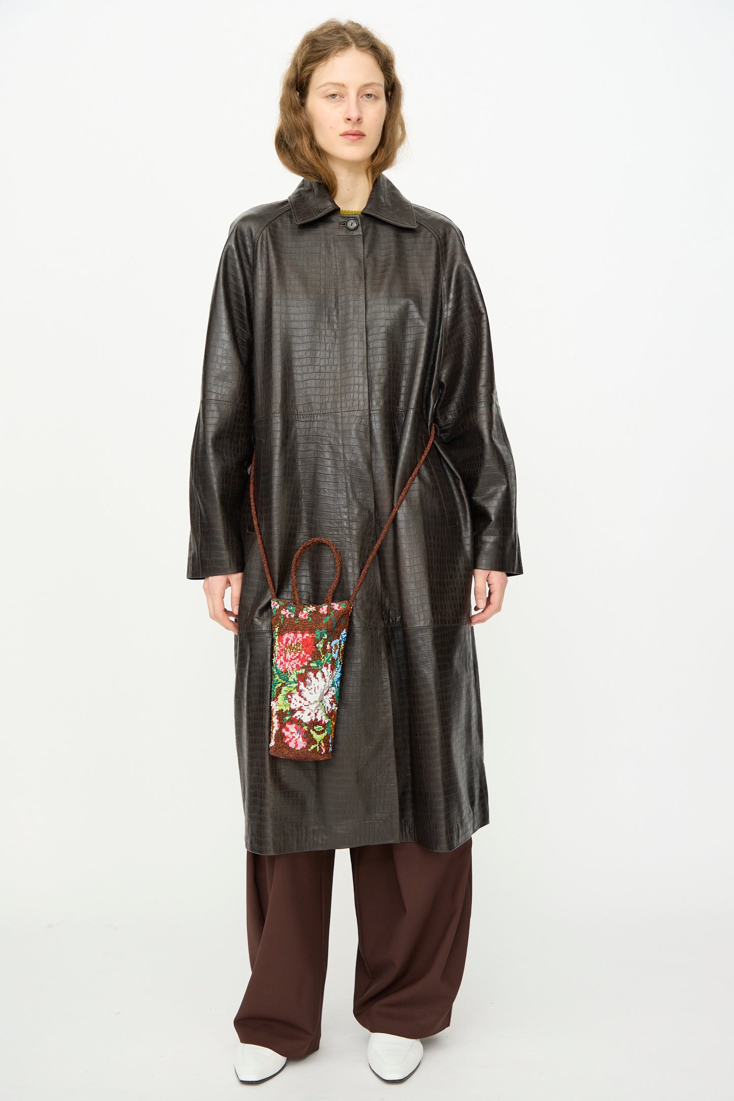 A person wears a textured dark brown long coat, brown wide-leg pants, white shoes, and carries Cordera's Handmade Beaded Bag in Peonia, a small artisanal accessory crafted in Guatemala with a strap.