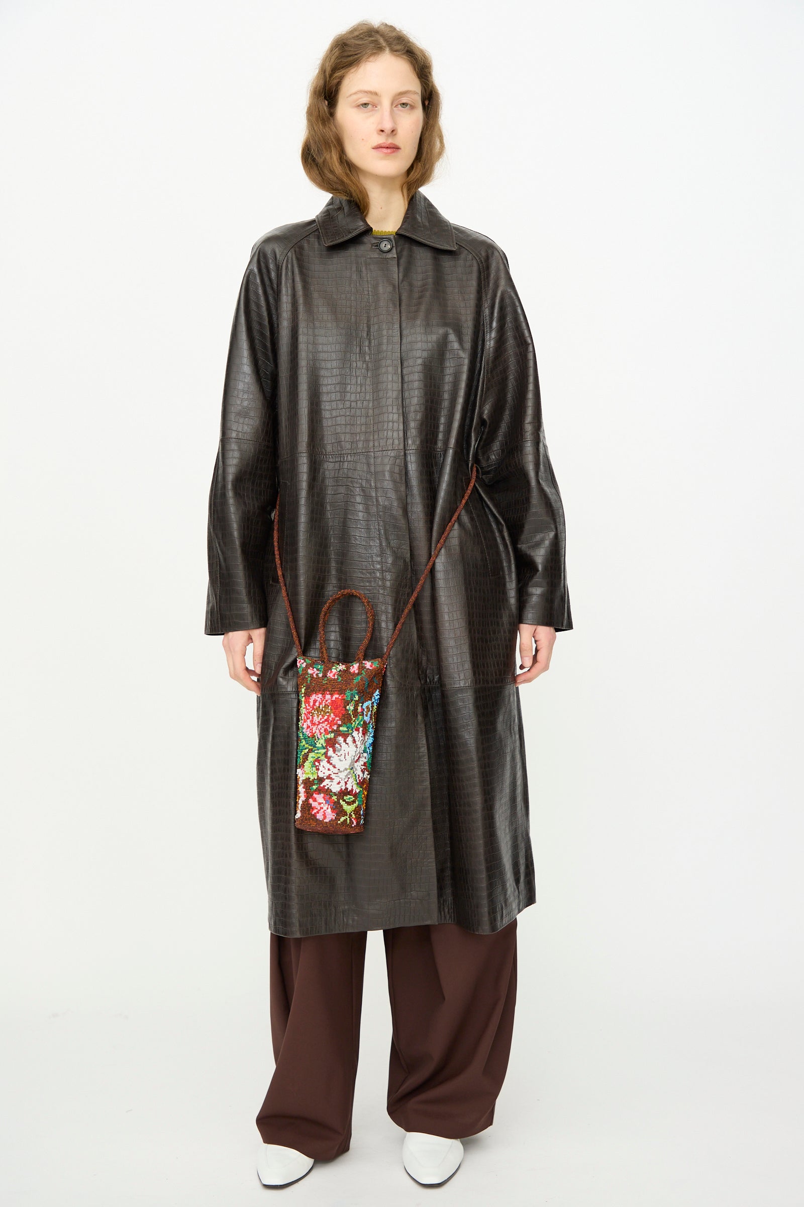 A person wears a textured dark brown long coat, brown wide-leg pants, white shoes, and carries Cordera's Handmade Beaded Bag in Peonia, a small artisanal accessory crafted in Guatemala with a strap.