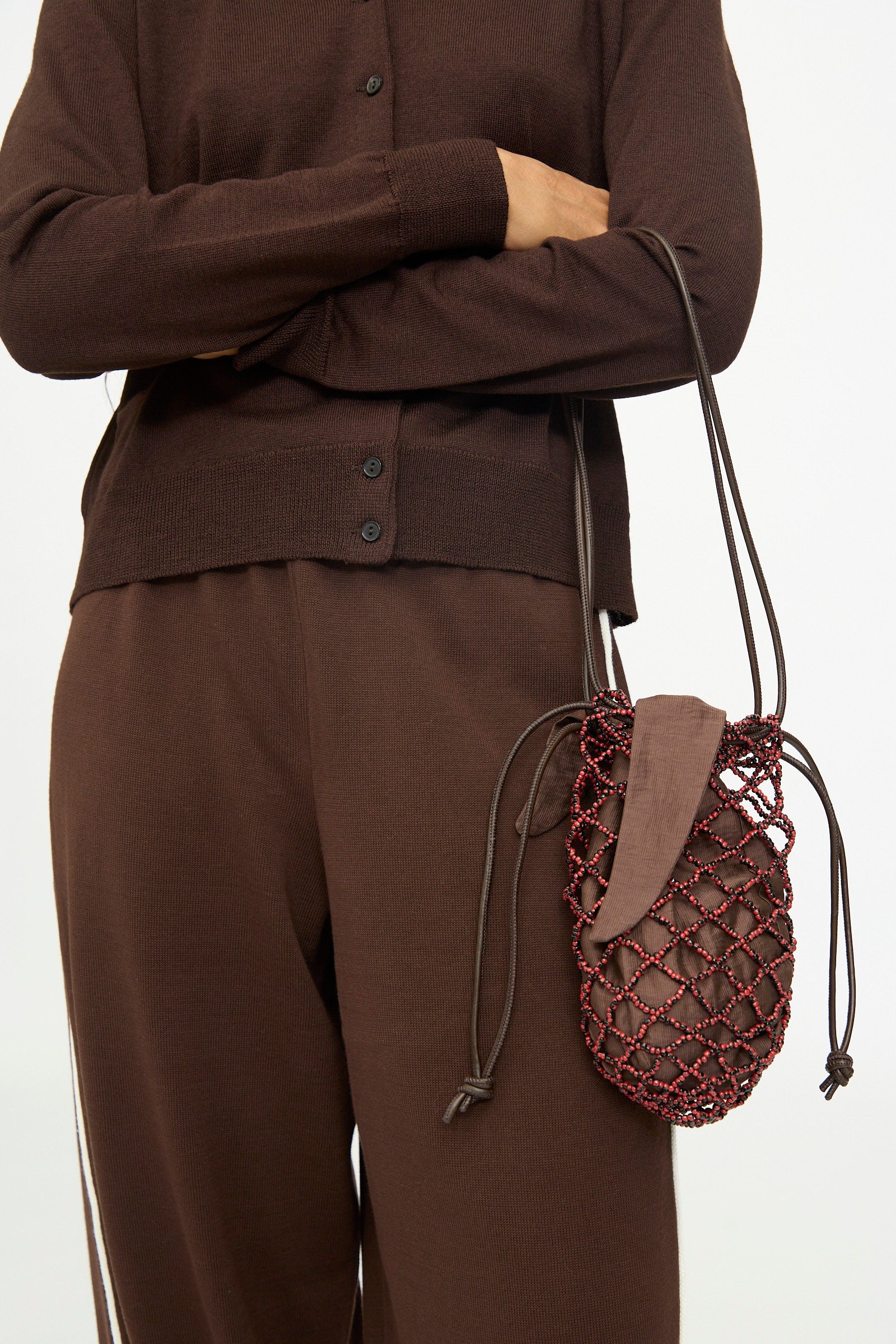 A person wearing a brown buttoned top and pants is holding the Cordera Handmade Beaded Net Bag, a small brown beaded handbag with a red beaded net design and drawstring straps, handcrafted in Guatemala.