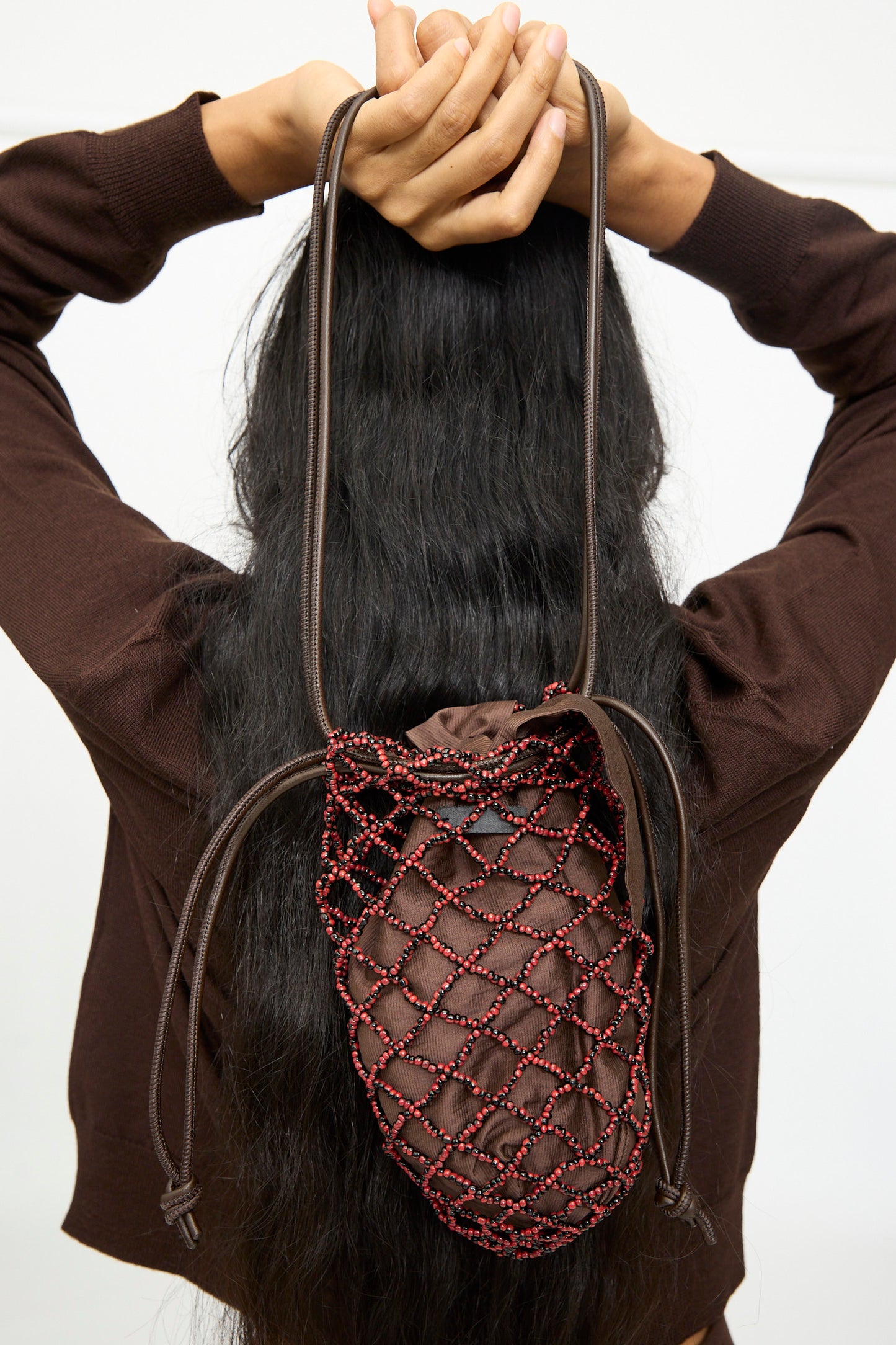 A person with long black hair wearing a brown sweater is holding their arms above their head and carrying the Cordera Handmade Beaded Net Bag—handcrafted in Guatemala—on their back.