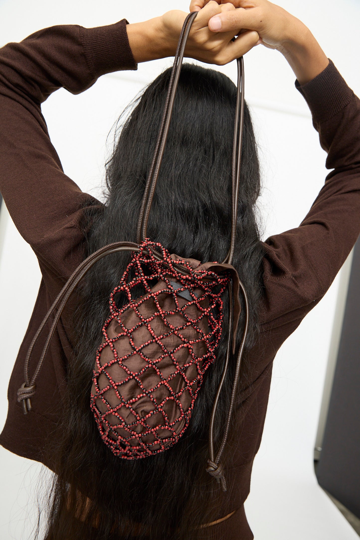 A person with long black hair wears a brown top and carries the Cordera Handmade Beaded Net Bag—handcrafted in Guatemala in red and brown—on their back, arms raised with hands behind their head.