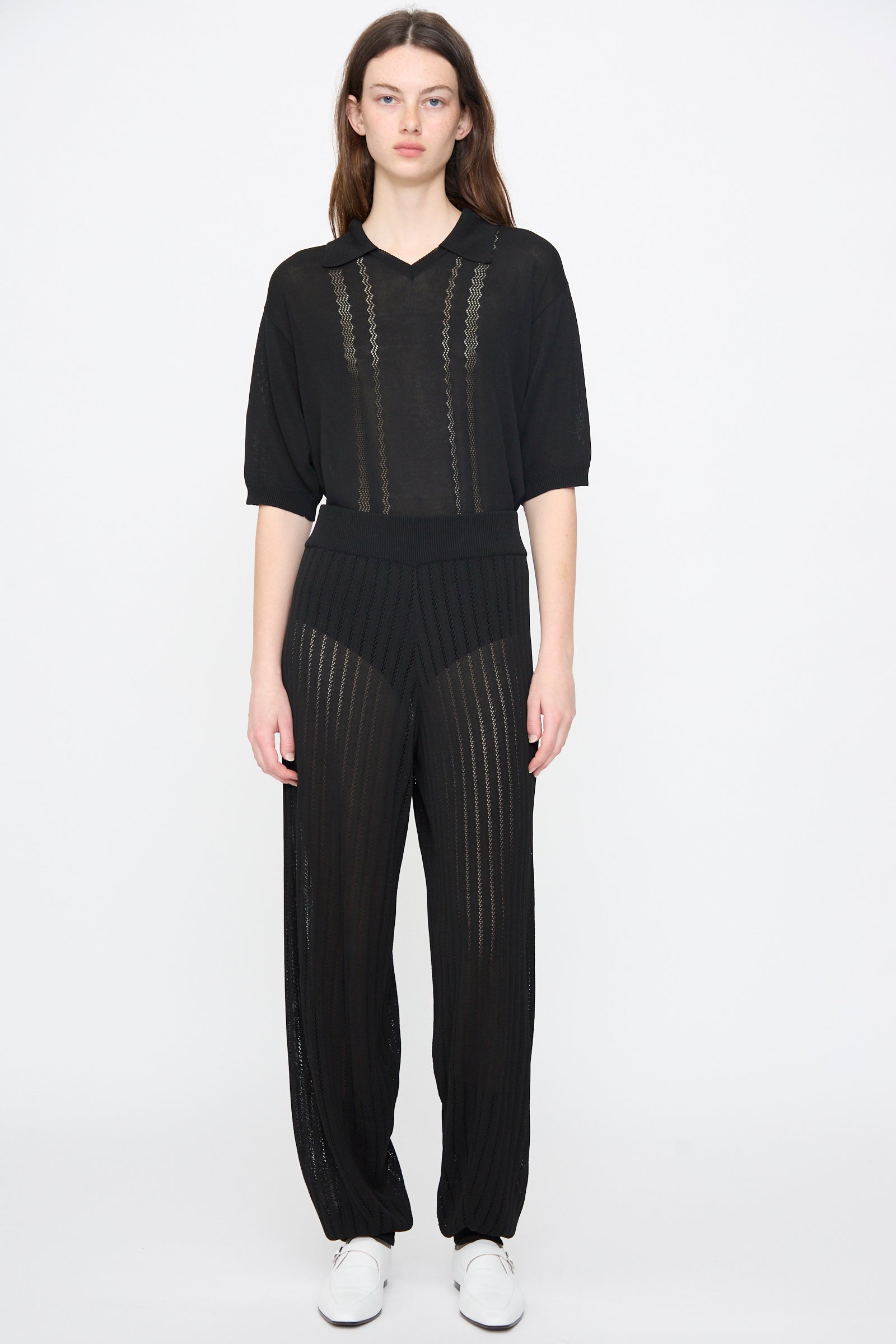A woman stands against a white background wearing Cordera's Lace Knit Pants in Black, styled with a sheer black top and white sneakers.