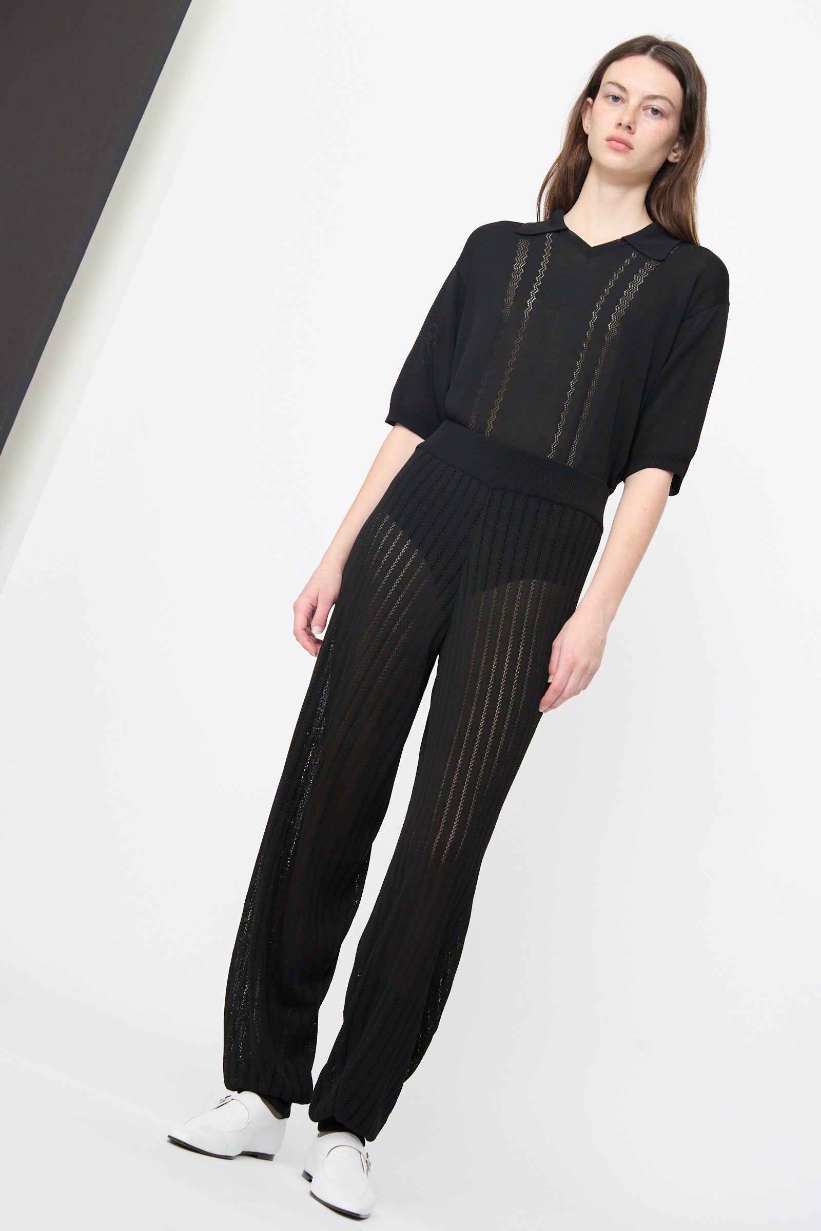 A woman stands against a white background wearing Cordera's Lace Knit Pants in Black, styled with a semi-sheer black top and white shoes.