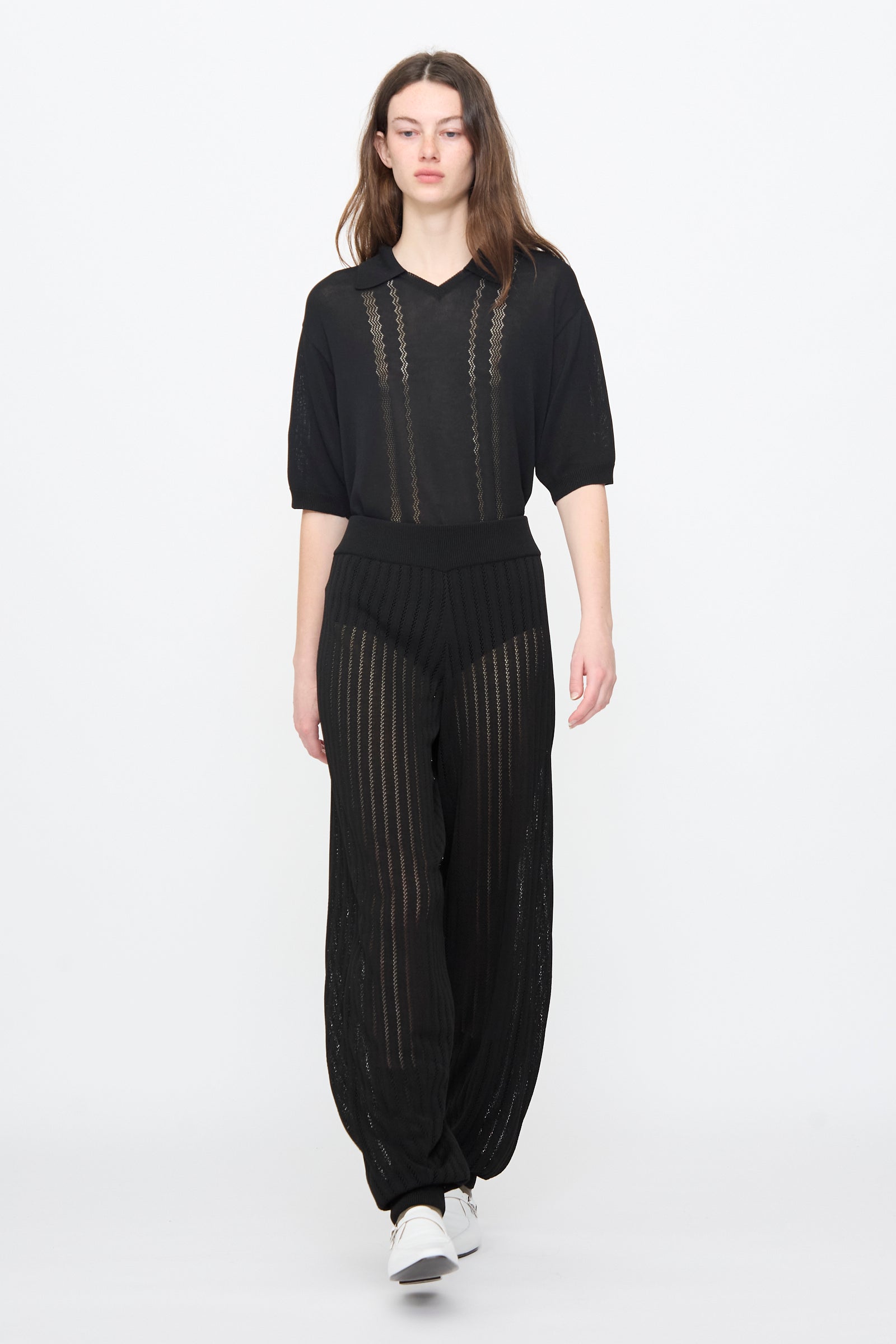 A woman stands against a plain white background wearing Cordera’s Lace Knit Pants in Black with a black sheer half-sleeve blouse and white sneakers.