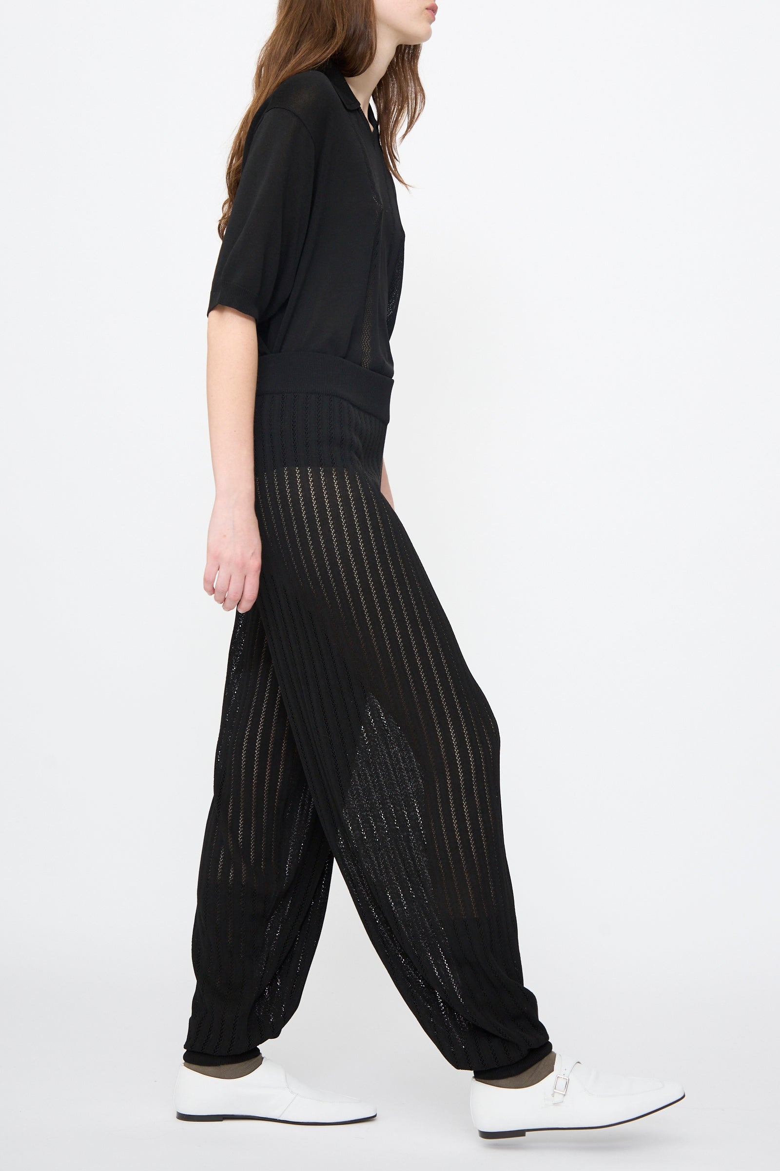 A person walks in profile wearing Cordera’s Lace Knit Pants in Black with a black sheer top and white shoes, set against a plain white background.