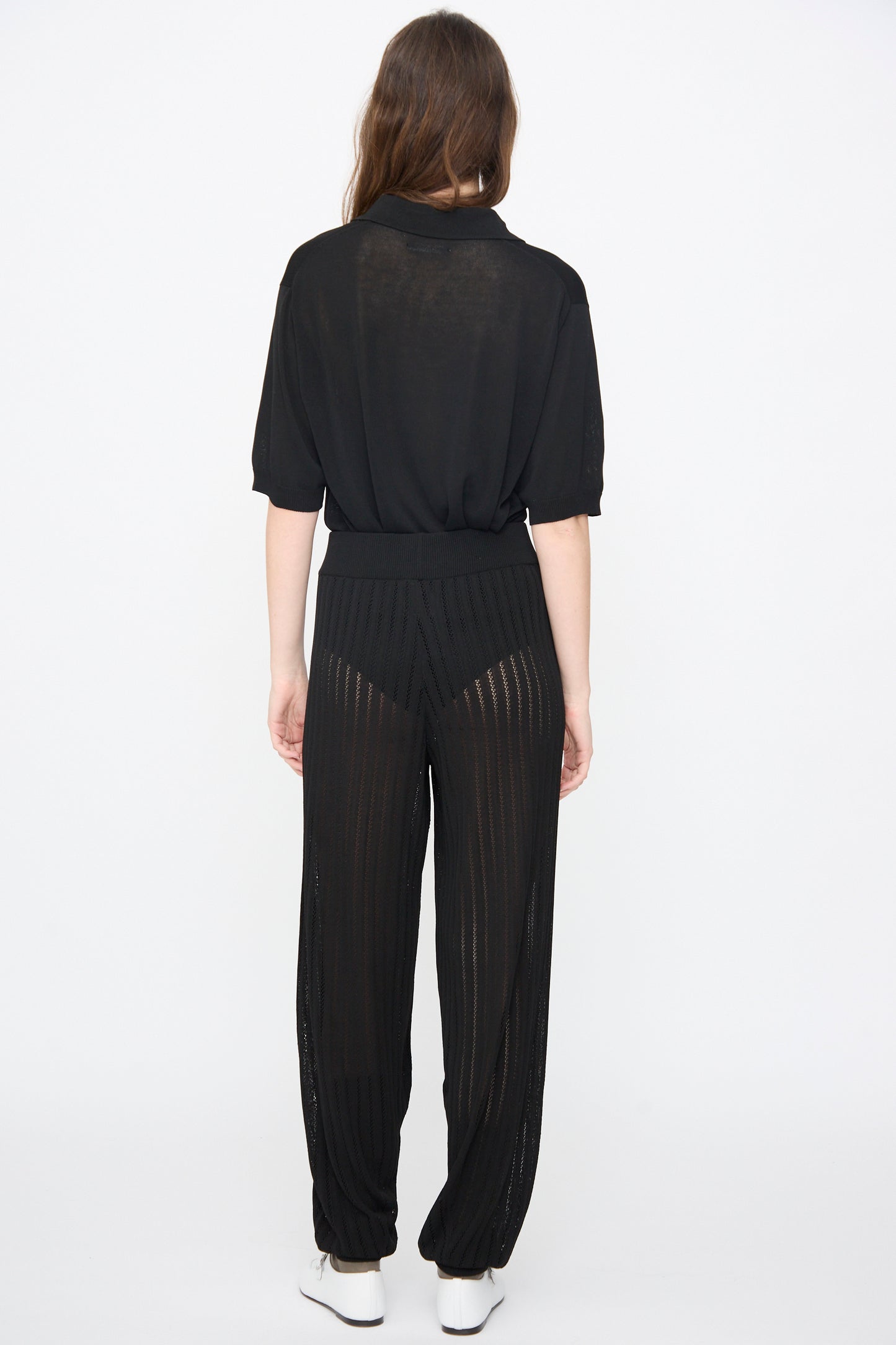 A woman stands facing away in Cordera's Lace Knit Pants in Black—a relaxed fit with sheer, striped details and visible black undergarments—paired with a loose black top and white shoes against a plain white background.