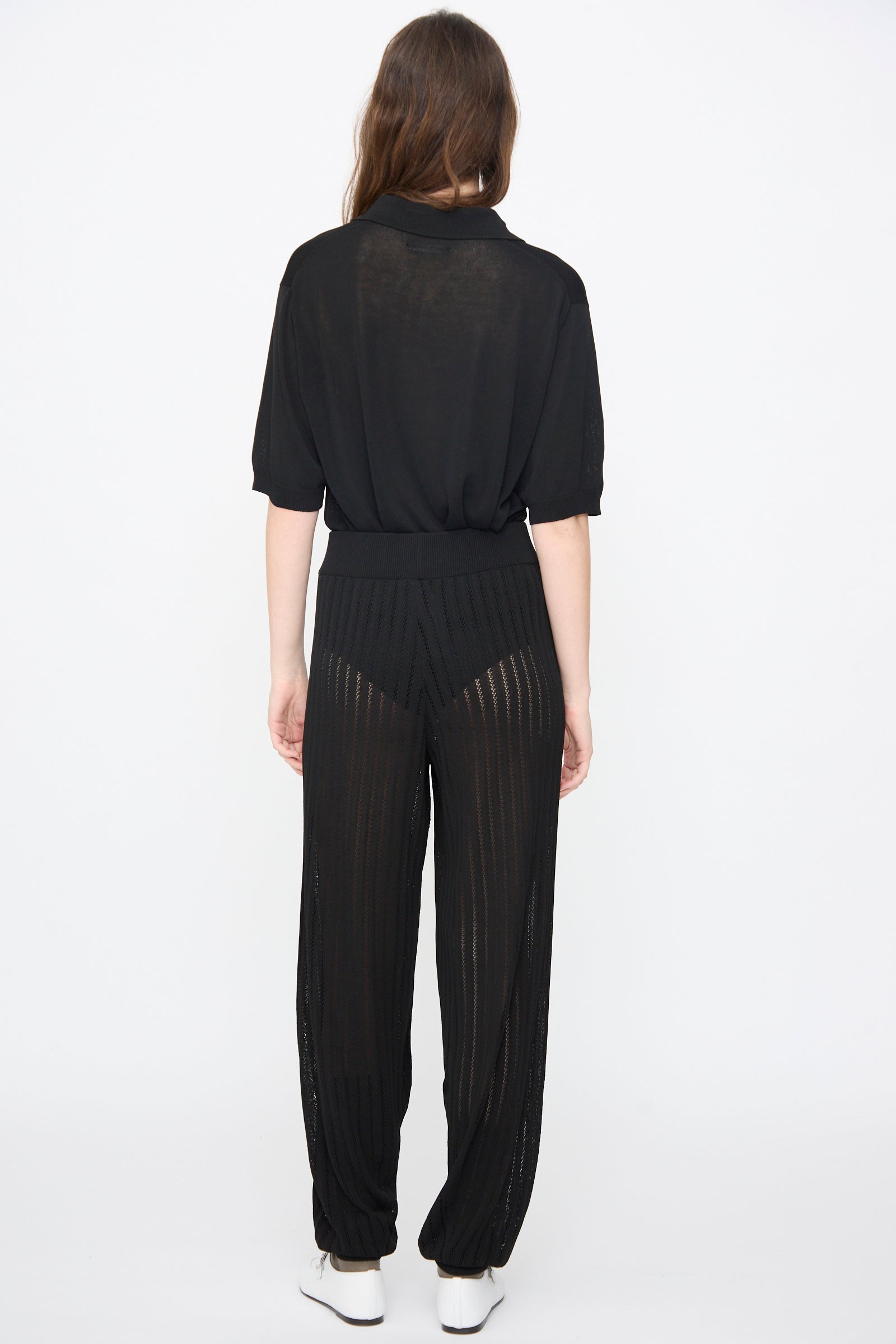 A woman stands facing away in Cordera's Lace Knit Pants in Black—a relaxed fit with sheer, striped details and visible black undergarments—paired with a loose black top and white shoes against a plain white background.