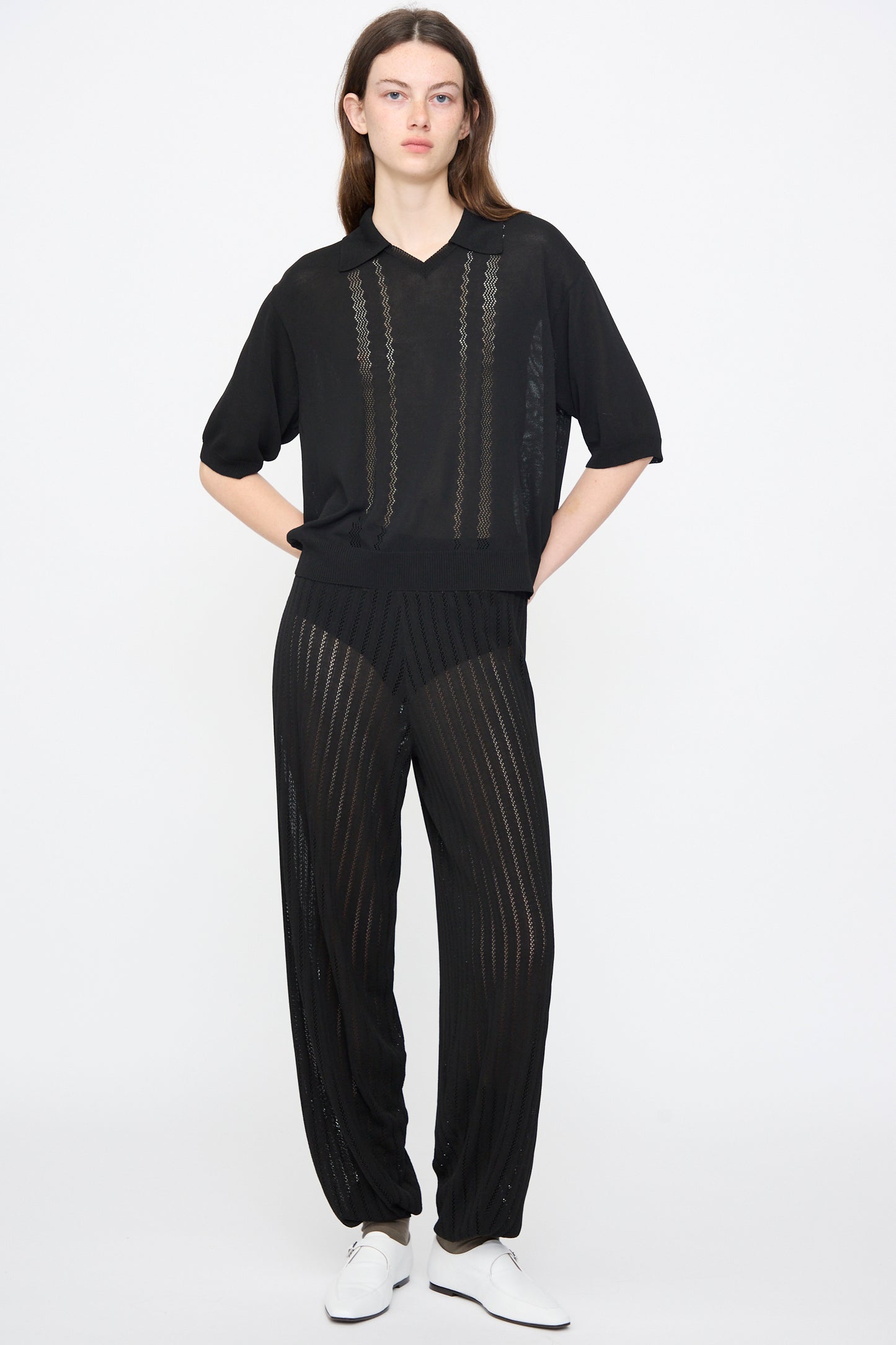 A person stands against a plain background wearing Cordera’s Lace Knit Pants in Black, styled with a loose black top and white shoes, hands in pockets and a neutral expression.