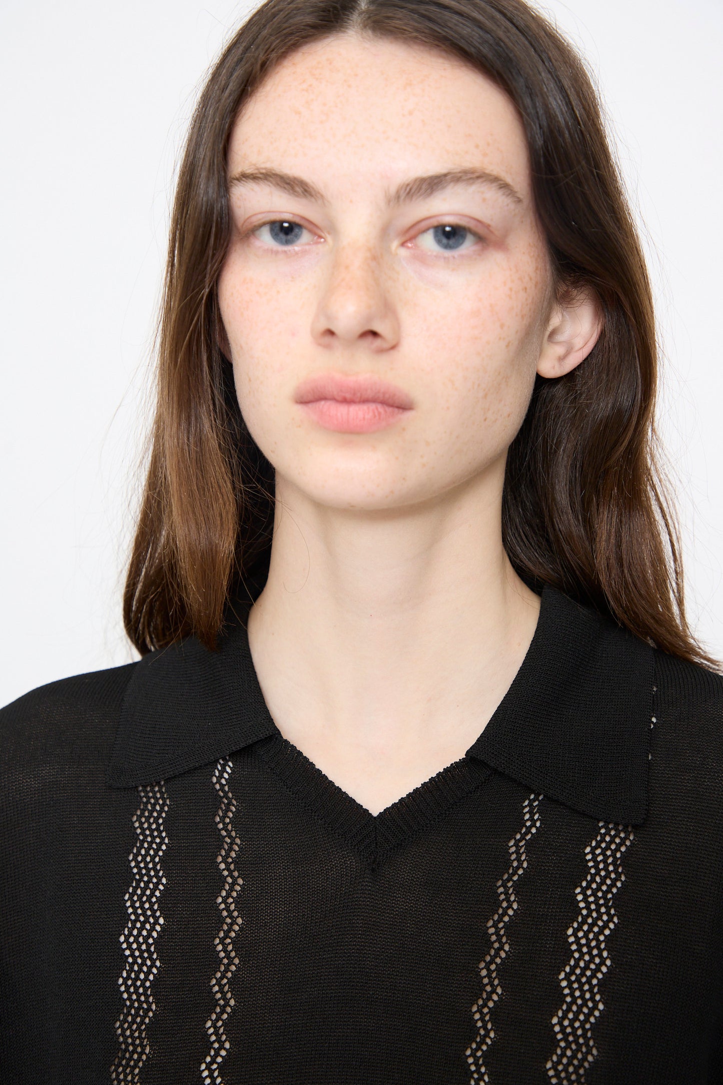 A woman with long brown hair and blue eyes models the Cordera Lace Knit Polo in Black, featuring lace design details and a collar, as she faces forward against a plain background.