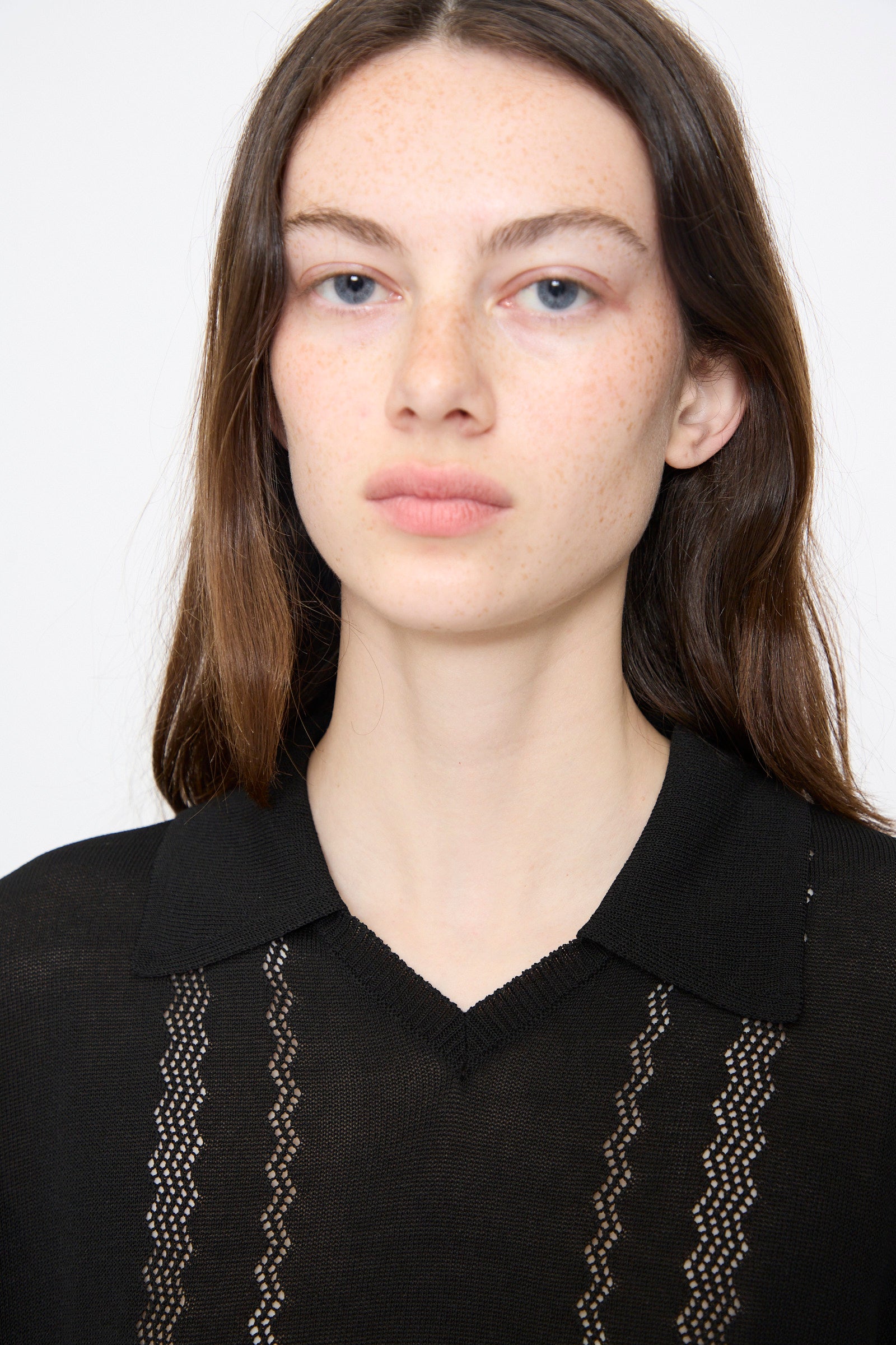 A woman with long brown hair and blue eyes models the Cordera Lace Knit Polo in Black, featuring lace design details and a collar, as she faces forward against a plain background.
