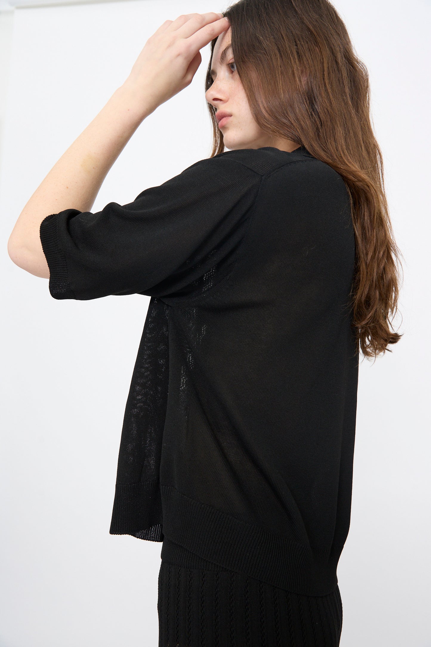 A person with long brown hair wears Cordera’s Lace Knit Polo in Black and faces sideways, raising one hand to their forehead against a plain background.