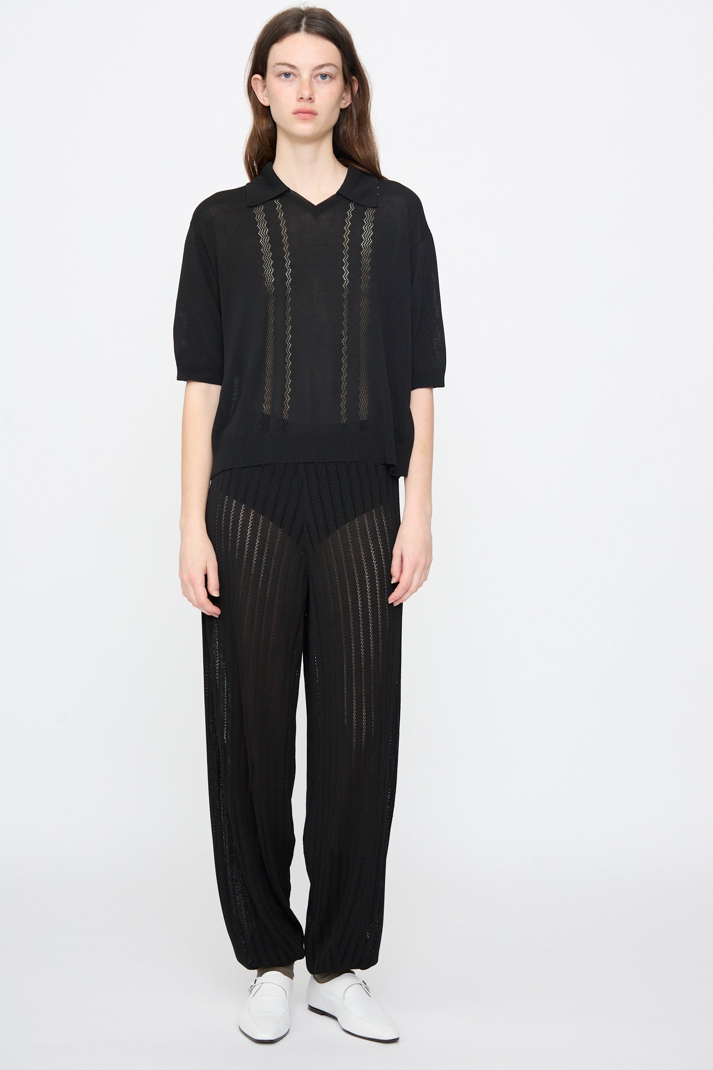 A person stands against a plain background wearing Cordera's Lace Knit Polo in Black with loose black sheer viscose pants and white shoes.