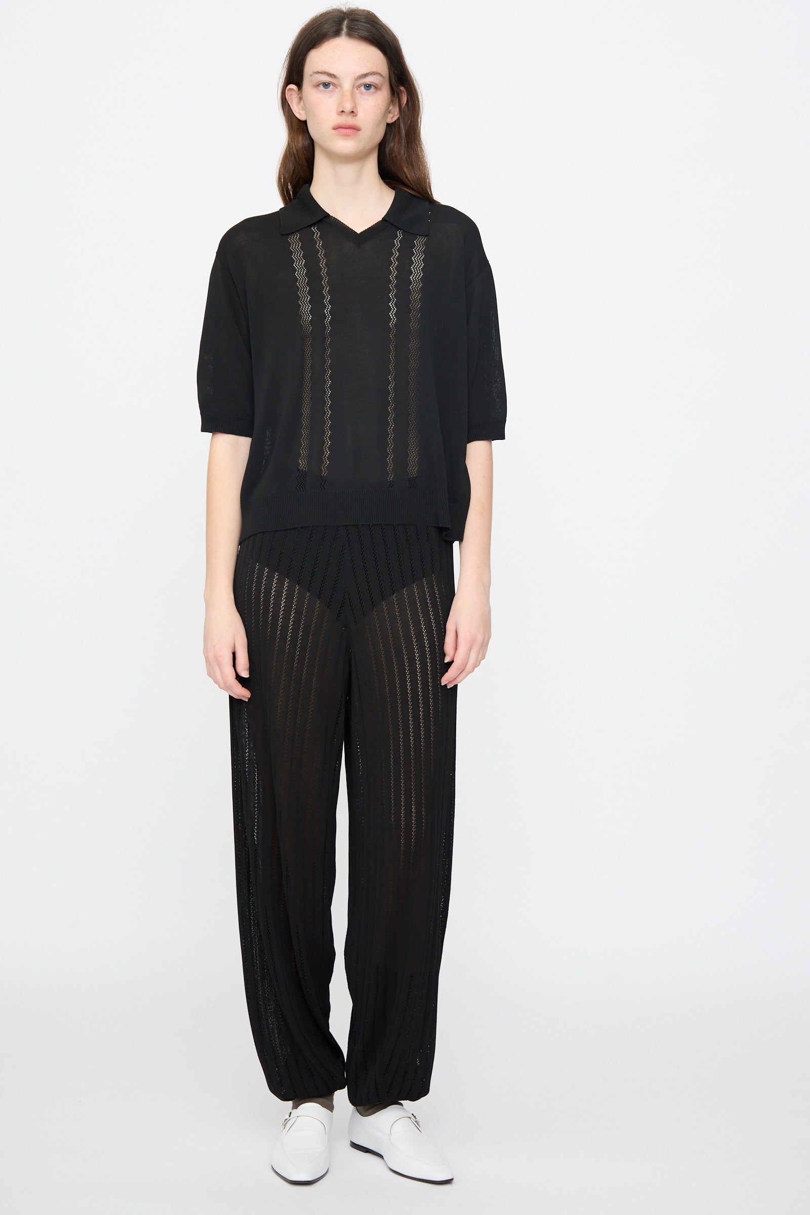 A person stands against a plain background wearing Cordera's Lace Knit Polo in Black with loose black sheer viscose pants and white shoes.