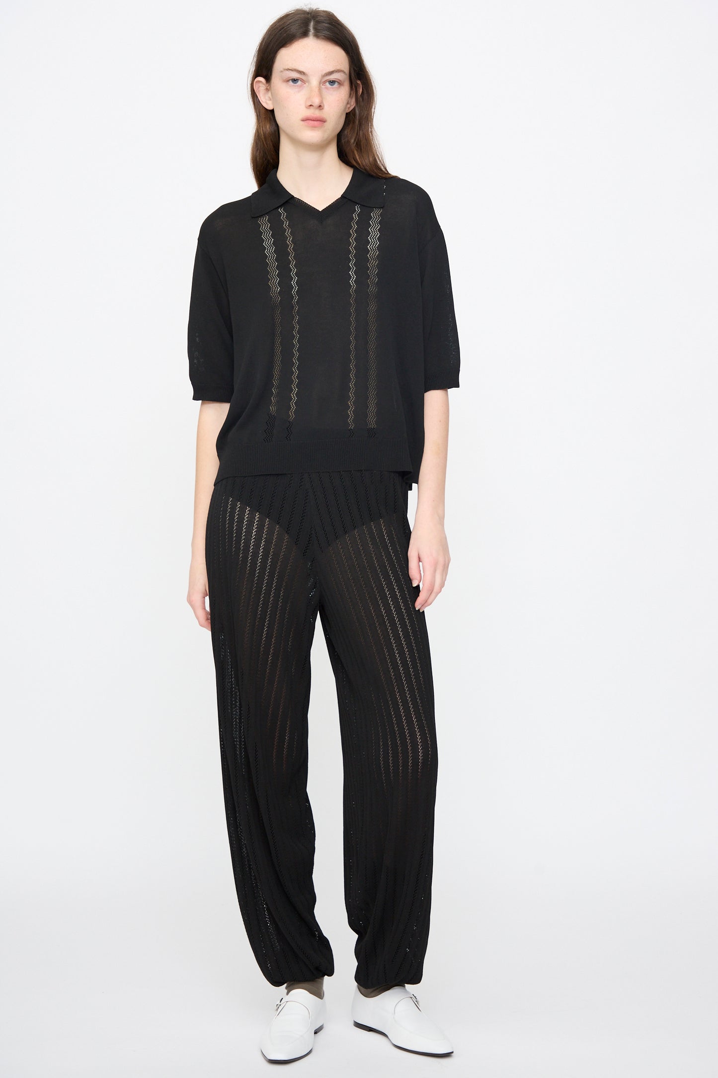 A person stands against a plain white background wearing the Cordera Lace Knit Polo in Black, featuring delicate lace details, paired with matching black pants and white shoes.
