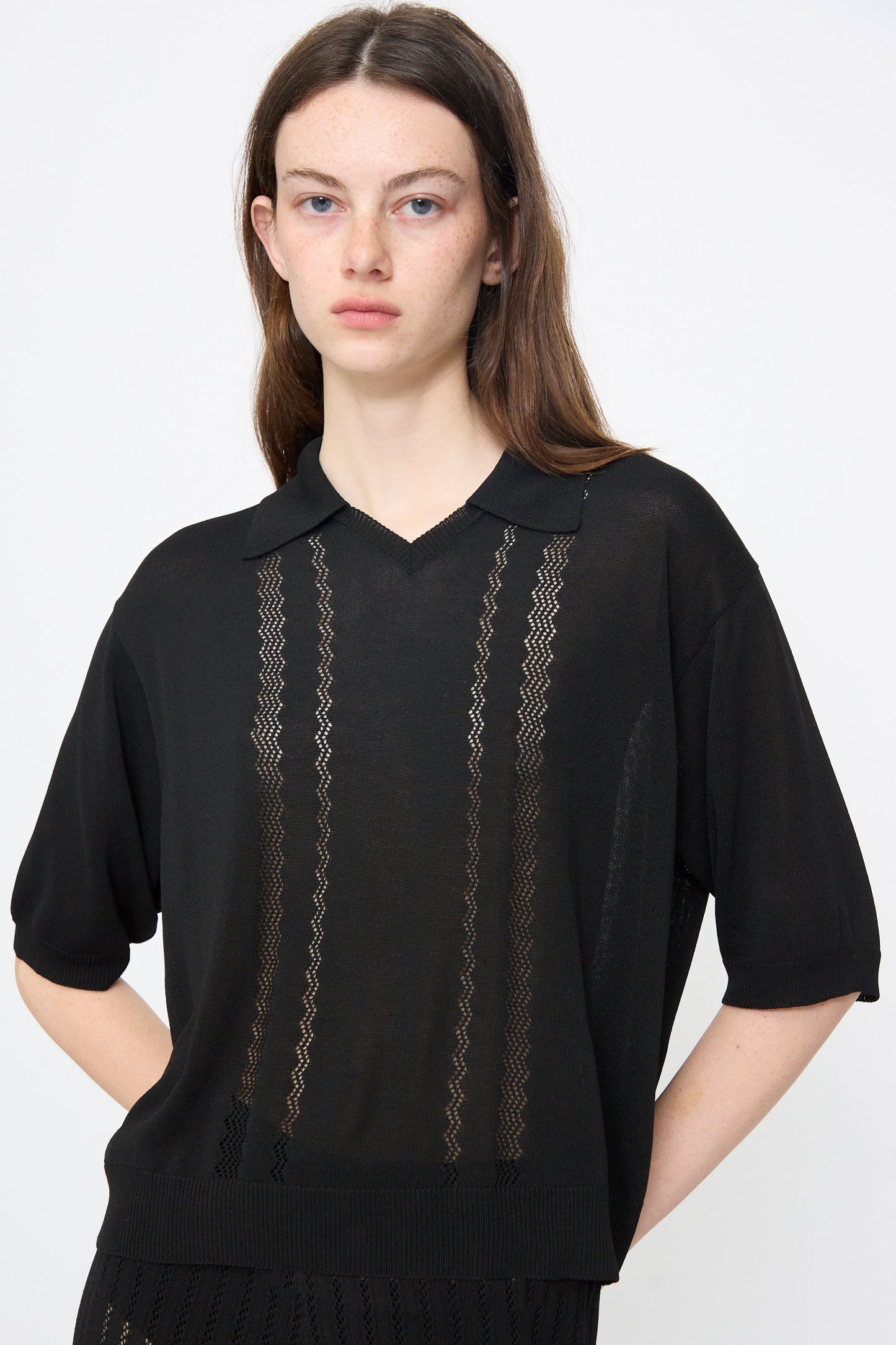A woman with long brown hair wears the Cordera Lace Knit Polo in Black, featuring short sleeves, a collar, and sheer zigzag-patterned viscose vertical stripes.