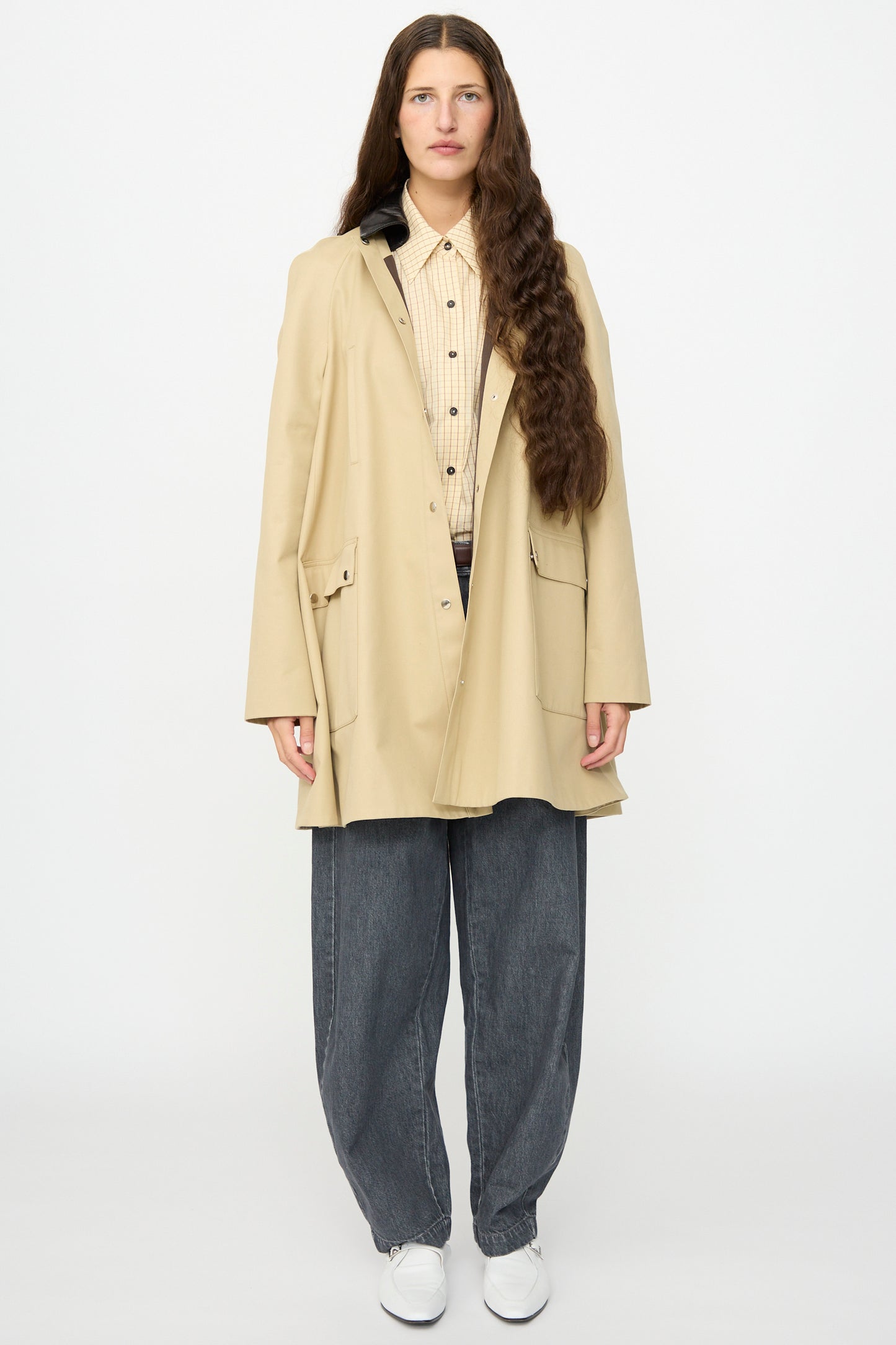 A person with long brown hair stands facing forward against a plain white background, wearing Cordera’s Leather Collar Voluminous Coat—a tan cotton A-line coat featuring a lambskin leather collar—layered over a patterned shirt, paired with loose gray jeans and white shoes.