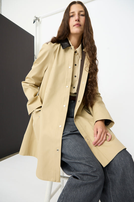 A person with long brown hair sits on a white stool in a minimalist studio, wearing Cordera’s Leather Collar Voluminous Coat, featuring a tan cotton A-line silhouette with a lambskin leather collar, layered over a checked shirt and wide-leg gray pants.