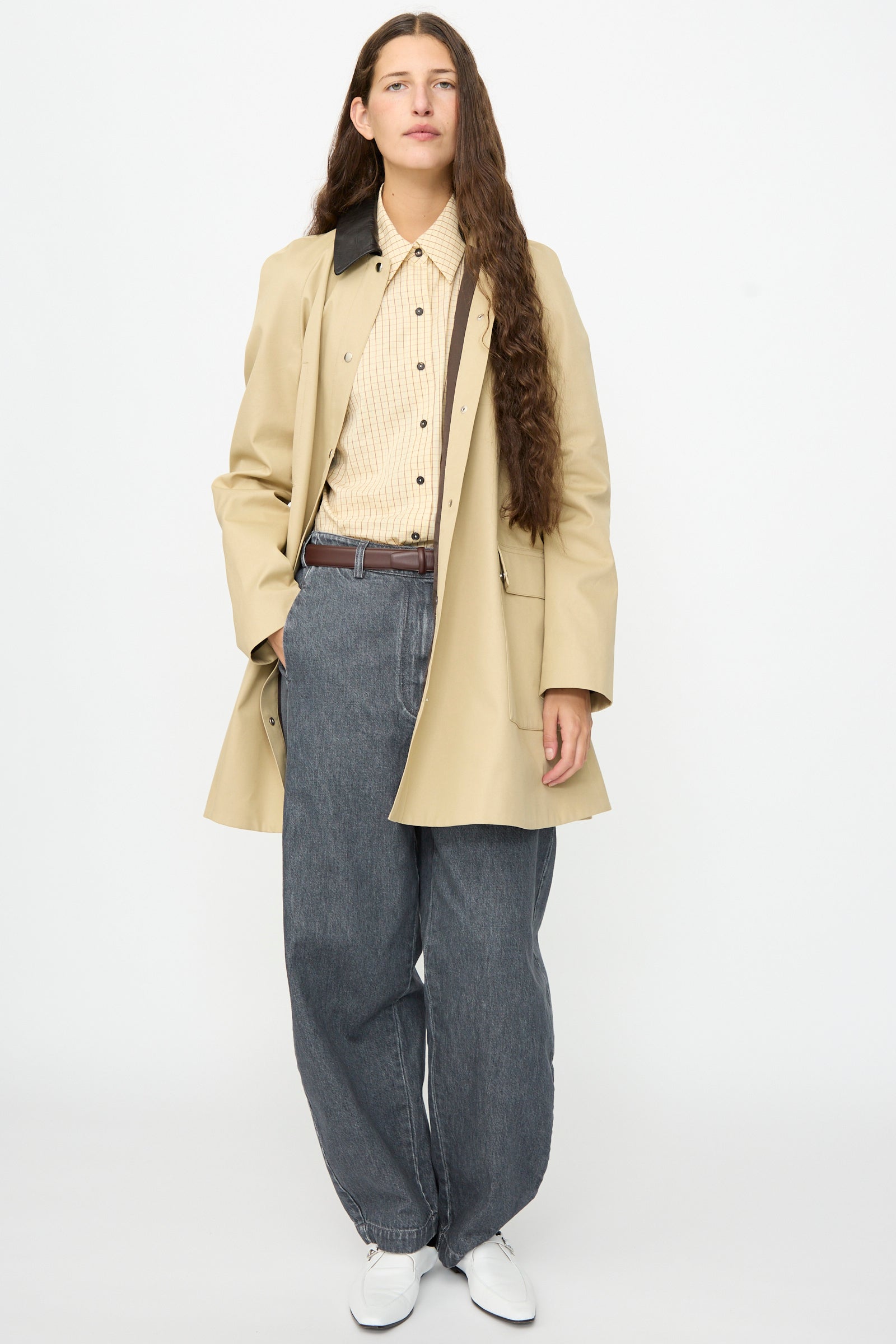 A person stands against a plain white background wearing the Cordera Leather Collar Voluminous Coat, featuring a tan cotton A-line silhouette with a lambskin leather collar, styled over a buttoned shirt, wide gray jeans, white shoes, and a brown belt.