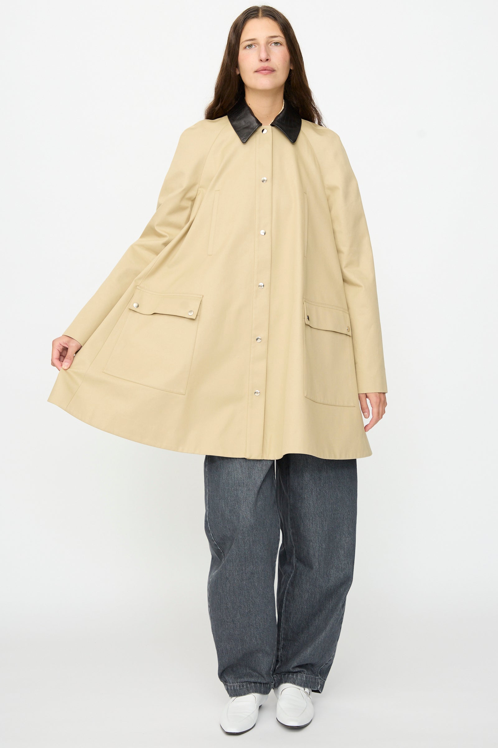 A person wearing Cordera’s Leather Collar Voluminous Coat in beige, which features a black lambskin leather collar, paired with gray pants and white shoes, stands against a plain white background.