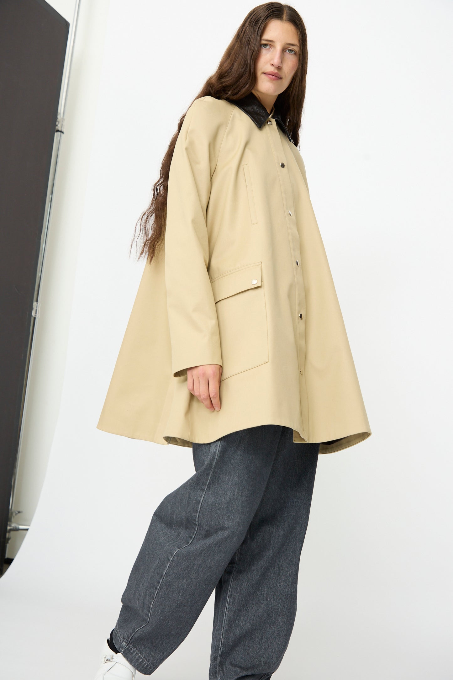 A person wearing the Cordera Leather Collar Voluminous Coat—featuring a tan cotton A-line silhouette with a lambskin leather collar—paired with dark gray jeans and white shoes, stands against a plain white background, looking at the camera.