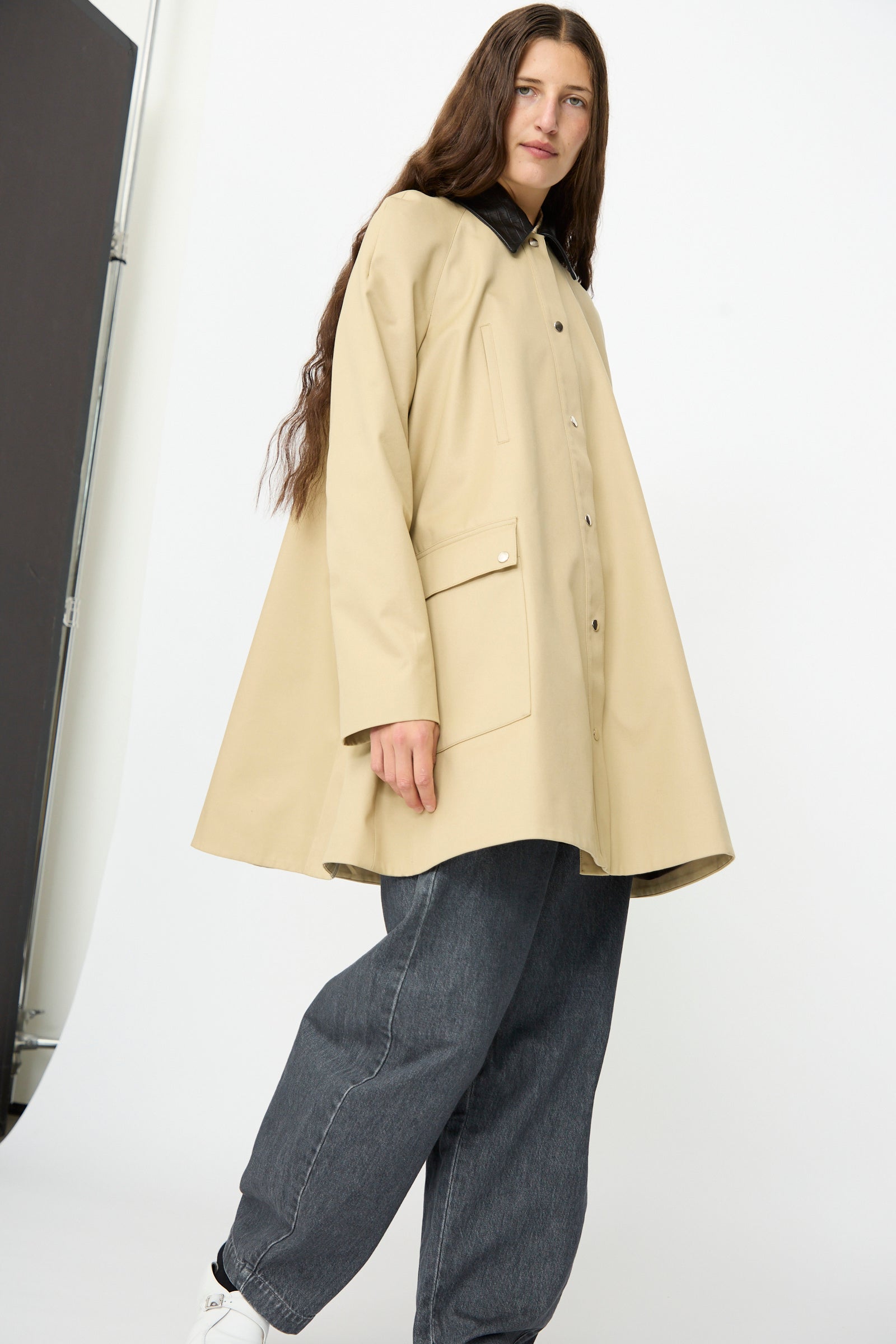 A person wearing the Cordera Leather Collar Voluminous Coat—featuring a tan cotton A-line silhouette with a lambskin leather collar—paired with dark gray jeans and white shoes, stands against a plain white background, looking at the camera.