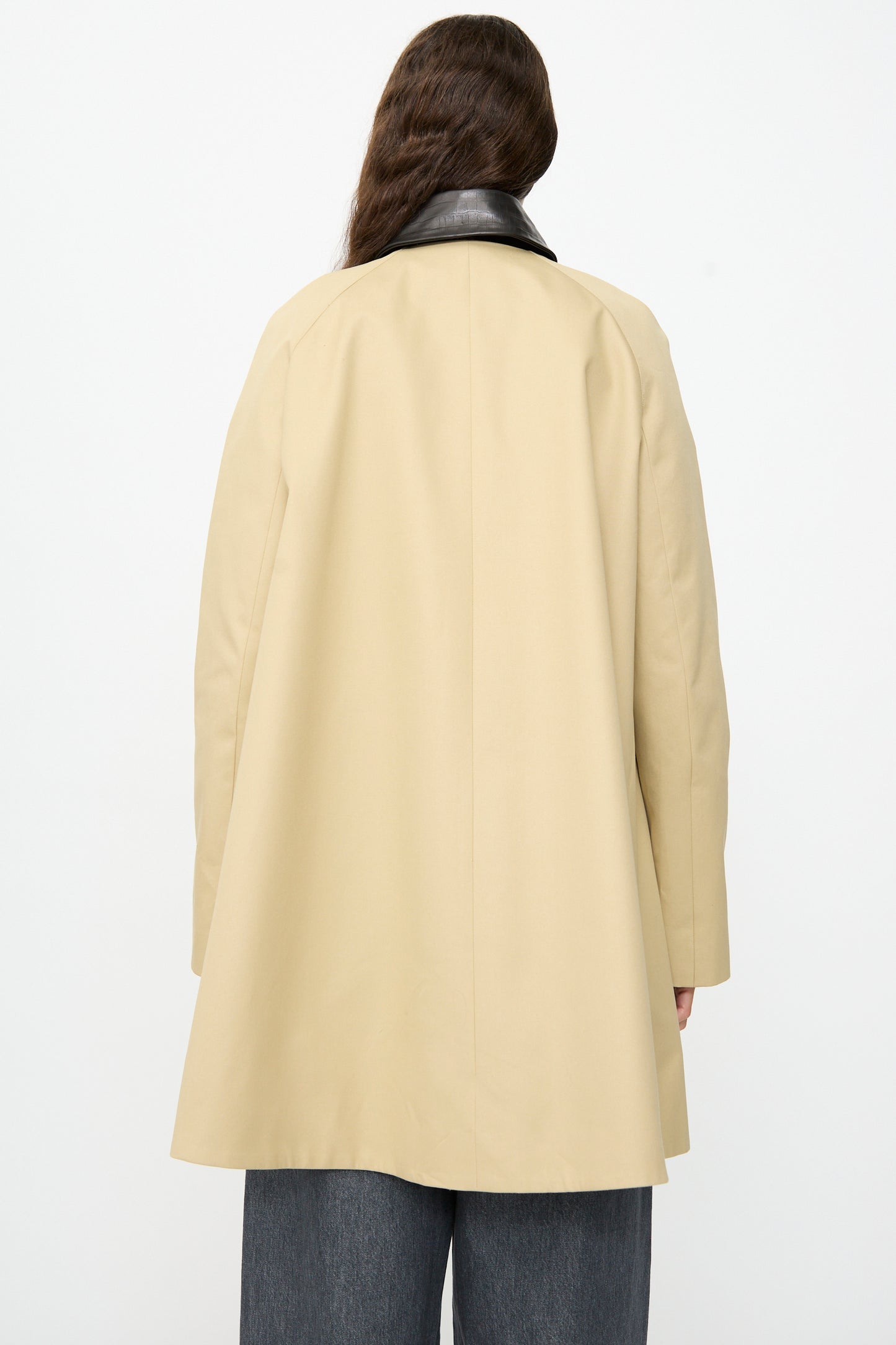 A person with long brown hair stands facing away, wearing the Cordera Leather Collar Voluminous Coat—an A-line tan cotton coat featuring a lambskin leather collar—and wide gray pants, set against a plain white background.