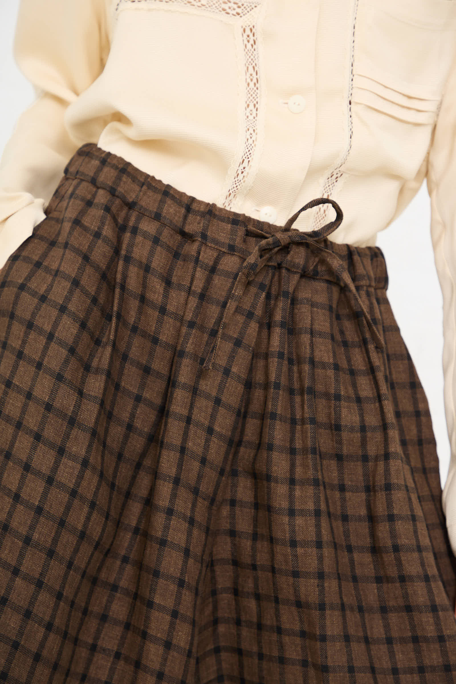 Close-up of a person wearing a cream-colored top with lace detail and the Linen Bermuda Short in Checkered by Cordera, featuring high-waisted, brown-checkered design with an elasticated waist and a drawstring bow at the front.