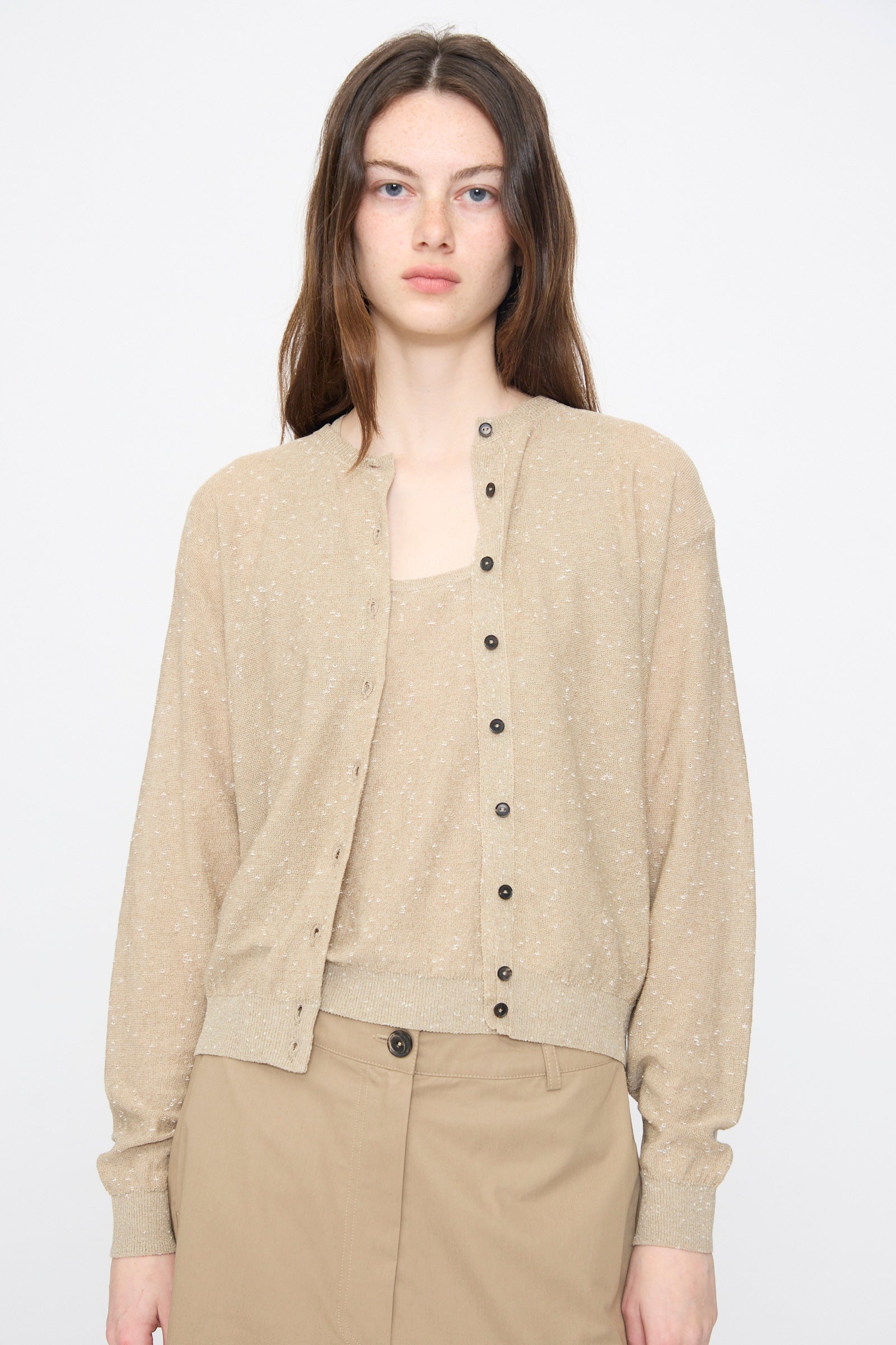 A woman with long brown hair models the Cordera Linen and Cotton Flecked Cardigan in Taupe, layered over a matching top and tan skirt, standing against a plain light gray background.