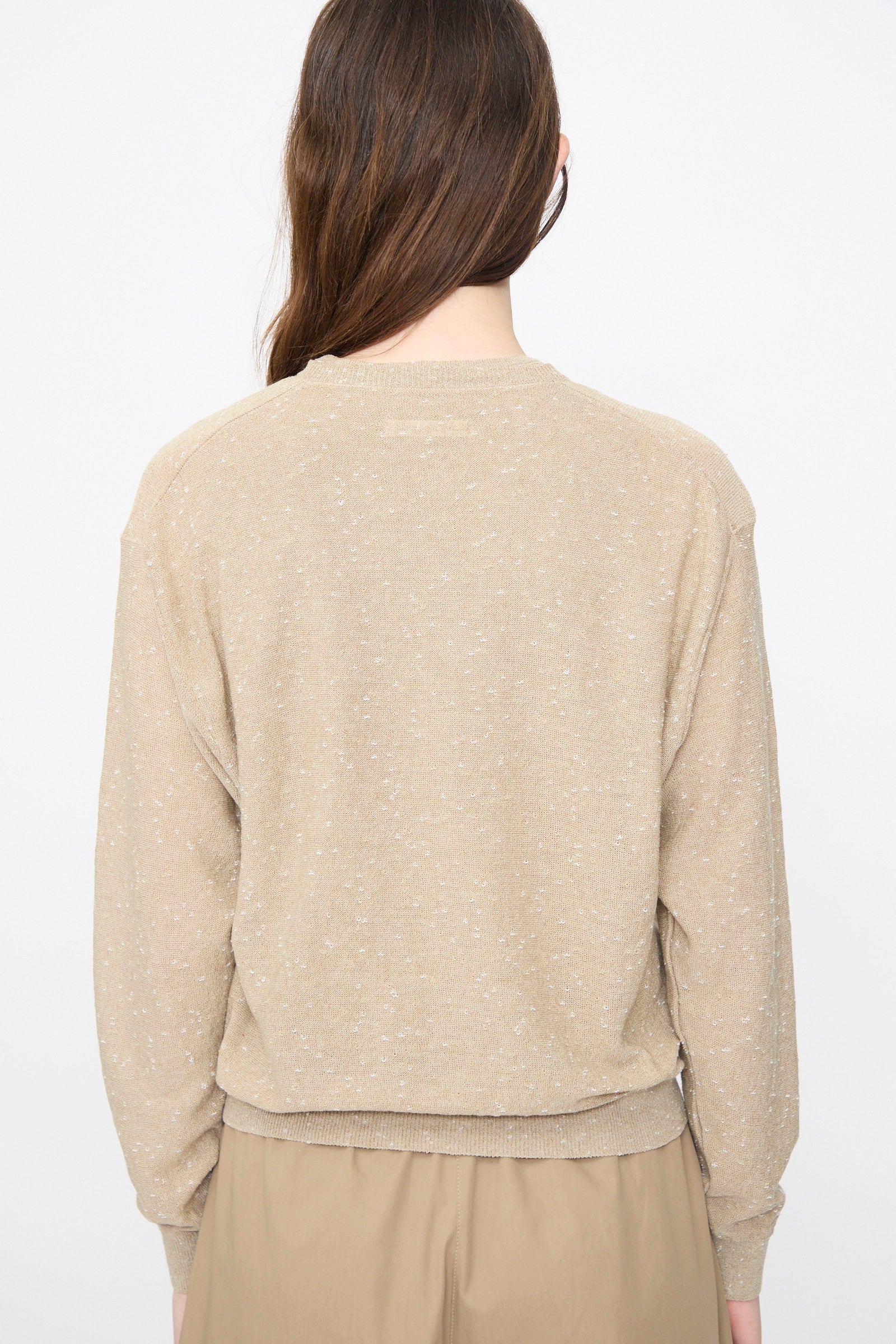 A person with long brown hair is seen from behind, wearing Cordera’s Linen and Cotton Flecked Cardigan in Taupe and light brown pants against a simple light background.
