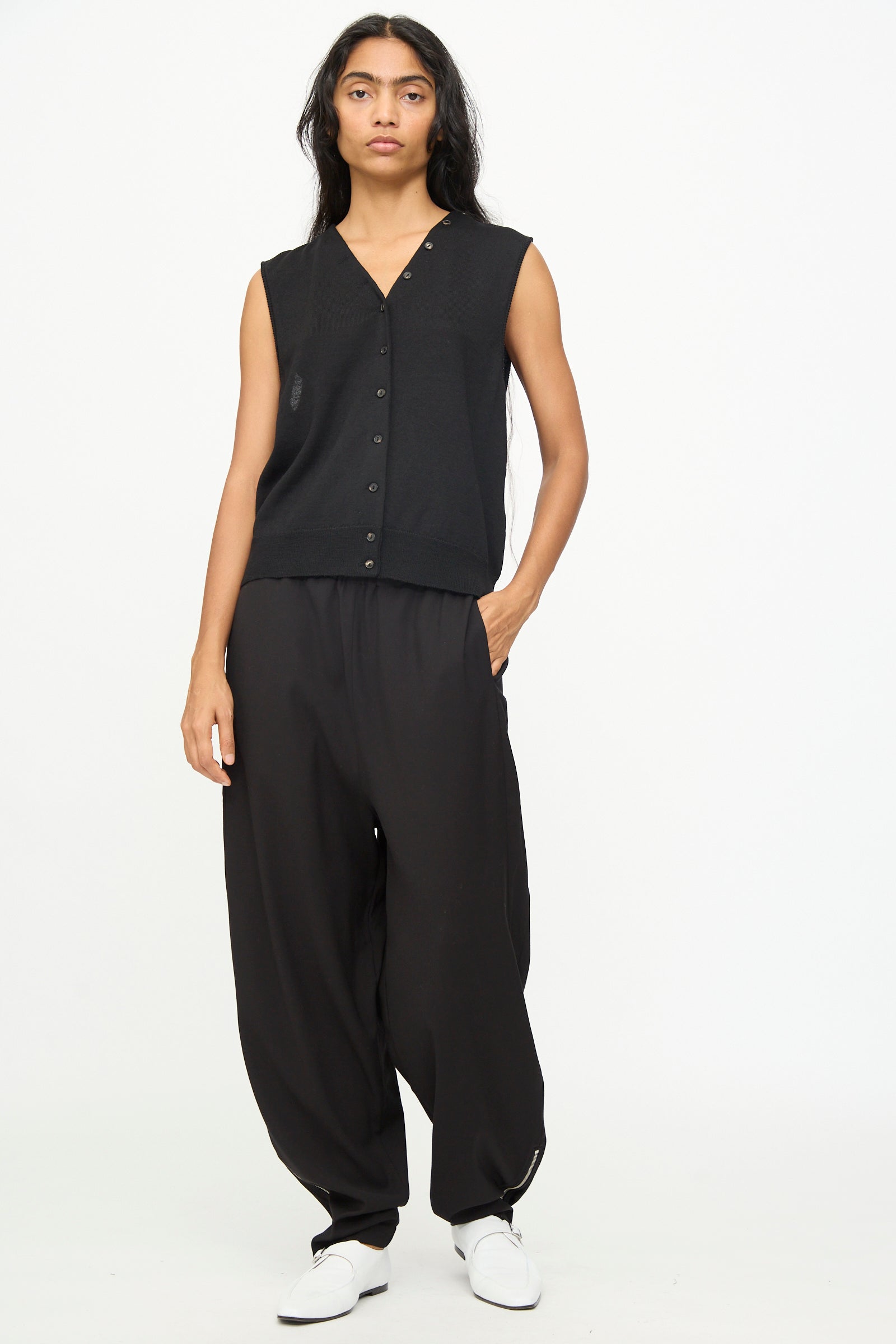 A person stands against a plain background, wearing Cordera Lounge Pants in Black—loose-fit lounge pants with an elastic waistband crafted from virgin wool—paired with a sleeveless black button-up vest and white shoes, hands by their sides and a neutral expression.