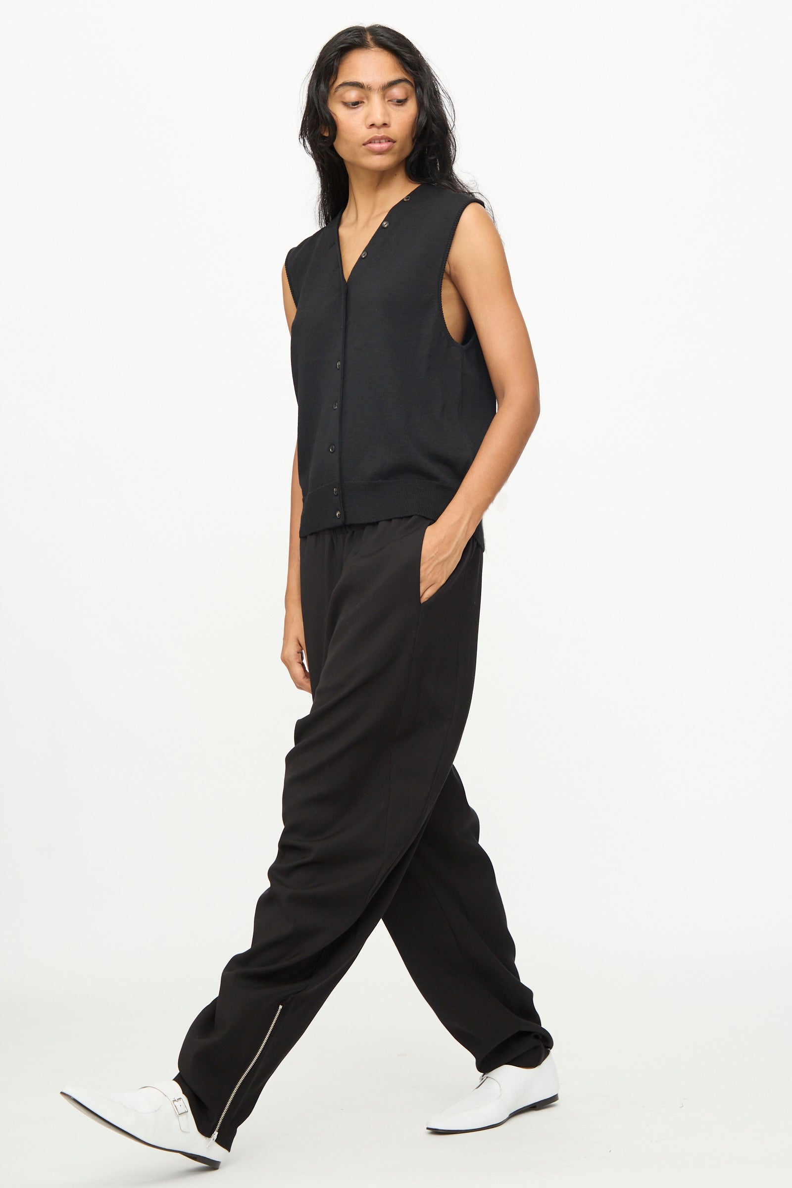 A person wearing Cordera's Lounge Pants in Black, paired with a black sleeveless buttoned top and white shoes, walks against a plain white background.