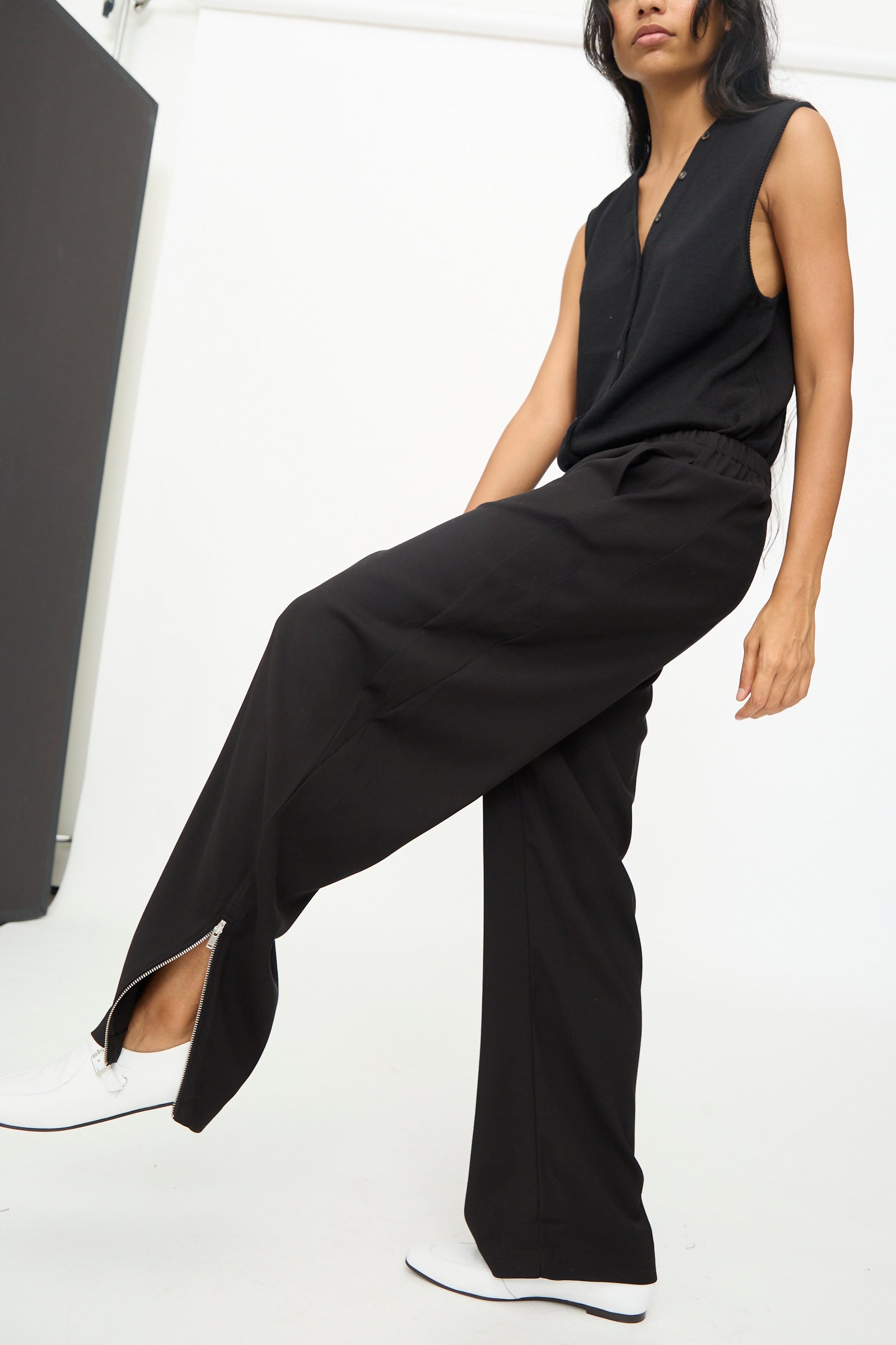 A person poses indoors against a minimal background, wearing Cordera's Lounge Pants in Black—wide-leg lounge pants crafted from virgin wool with an elastic waistband—paired with a sleeveless black top and white shoes. They lift one leg slightly.