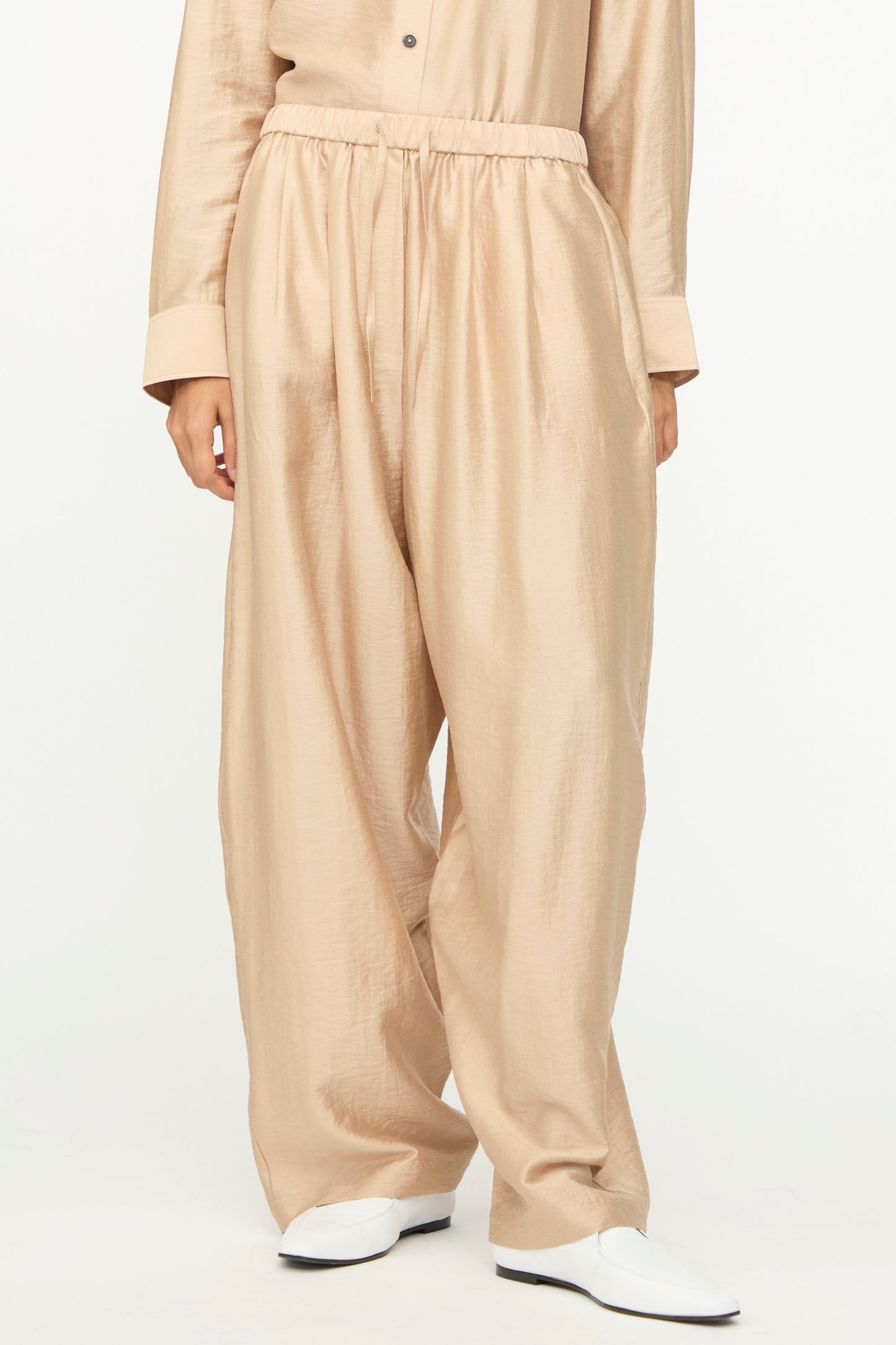A person wearing Cordera's Maxi Pants in Beige—loose, shiny wide-leg pants with an elastic waistband crafted from a soft lyocell blend—paired with a matching long-sleeve top and white shoes, standing against a plain white background.