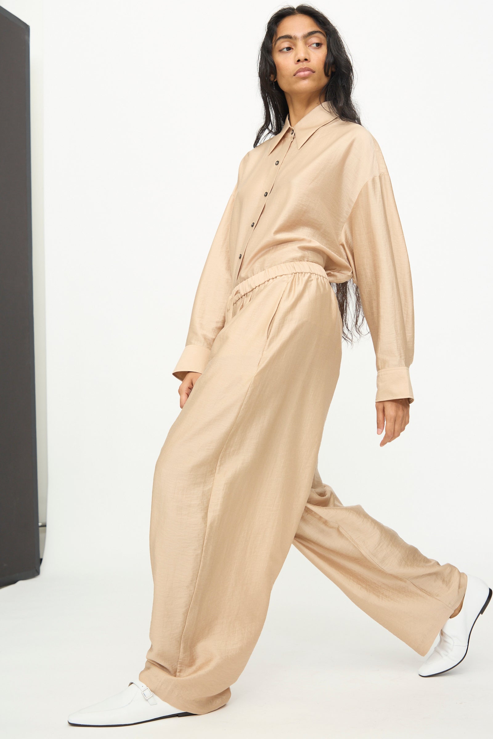 A person wearing Cordera's Maxi Pants in Beige—a loose, wide-leg lyocell blend style—paired with a matching beige button-up shirt and white shoes, stands against a plain white background.