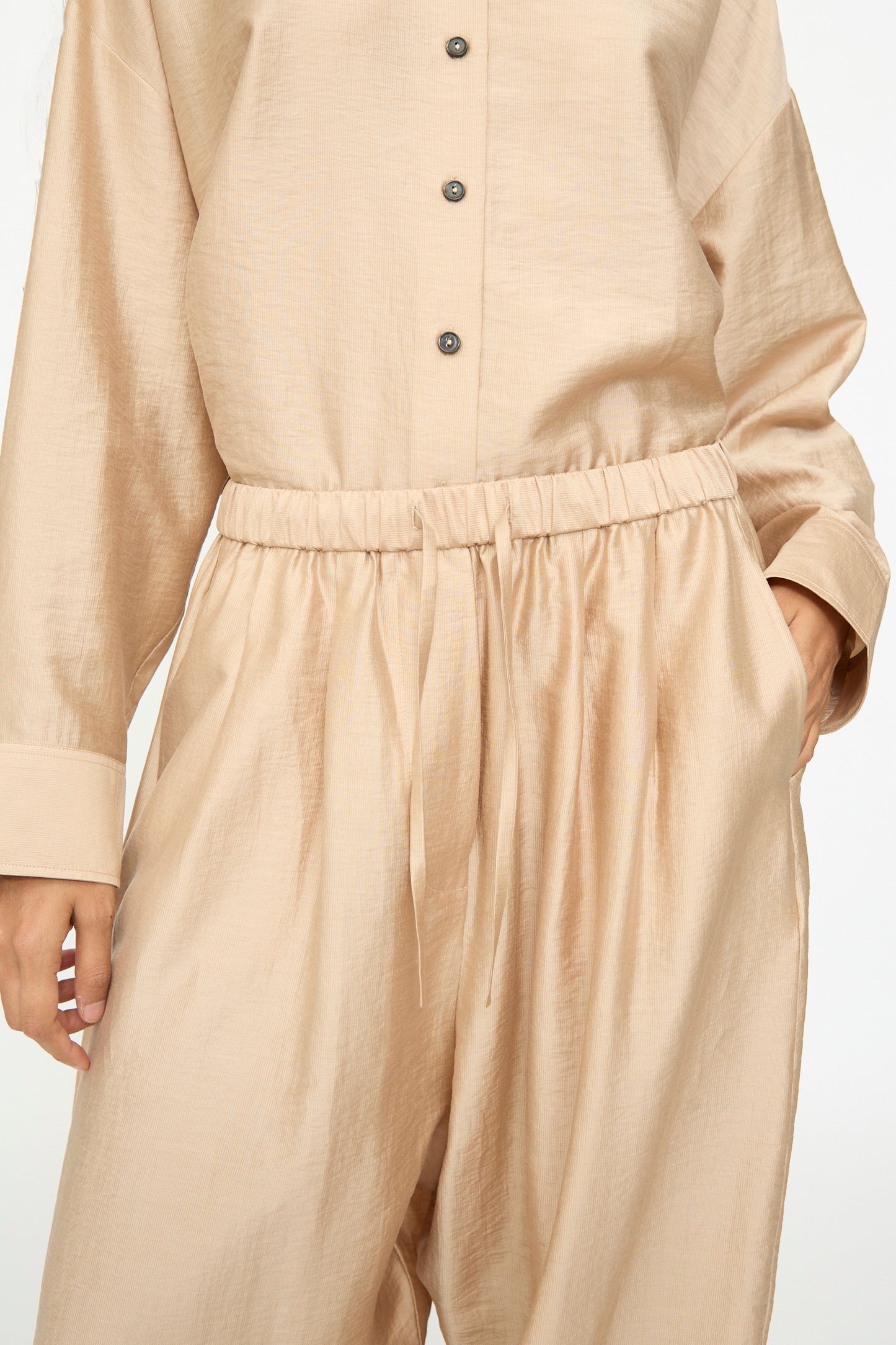 A person wearing Cordera’s Maxi Pants in Beige, styled with their hands in the pockets. The wide-leg drawstring pants and the loose, long-sleeve buttoned top are crafted from a soft lyocell blend for a relaxed, coordinated look.