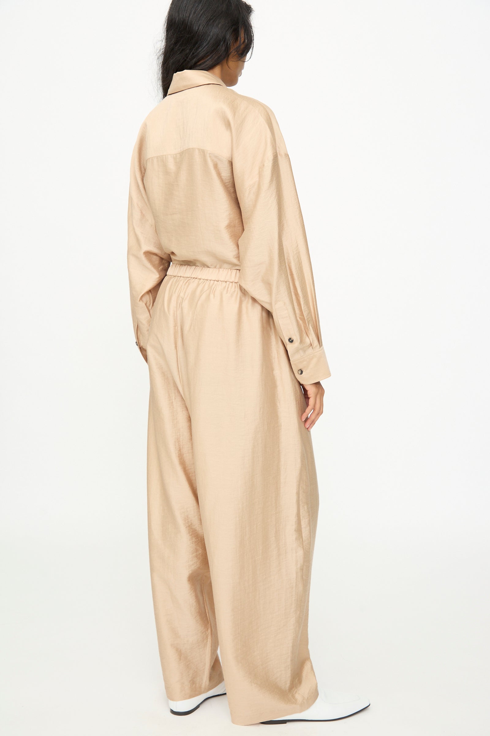 A person is seen from the back against a white background, wearing Cordera's Maxi Pants in Beige—a pair of wide-leg, lyocell blend pants—paired with a loose-fitting, tan long-sleeve shirt.