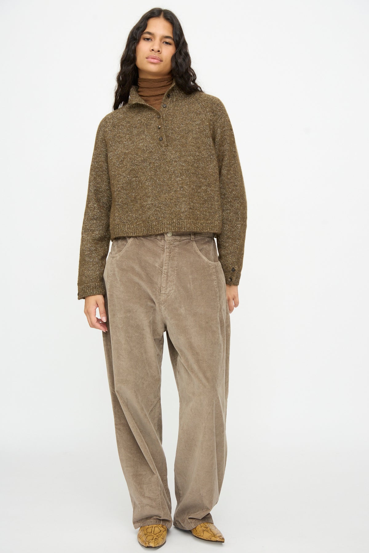 A person wears a Cordera Melange Polo Sweater in Pardo over a turtleneck and wide-leg trousers, styled with snakeskin-print ankle boots.