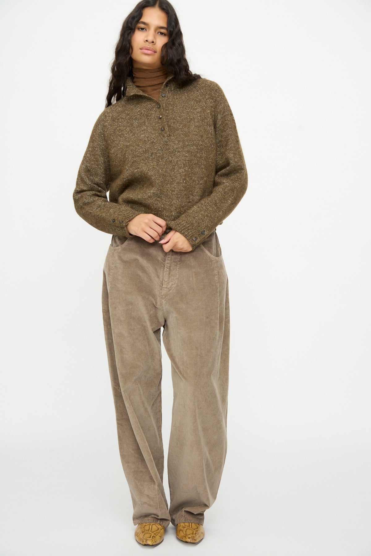 A person wears a brown, speckled Melange Polo Sweater in Pardo over a brown turtleneck and wide-leg trousers.