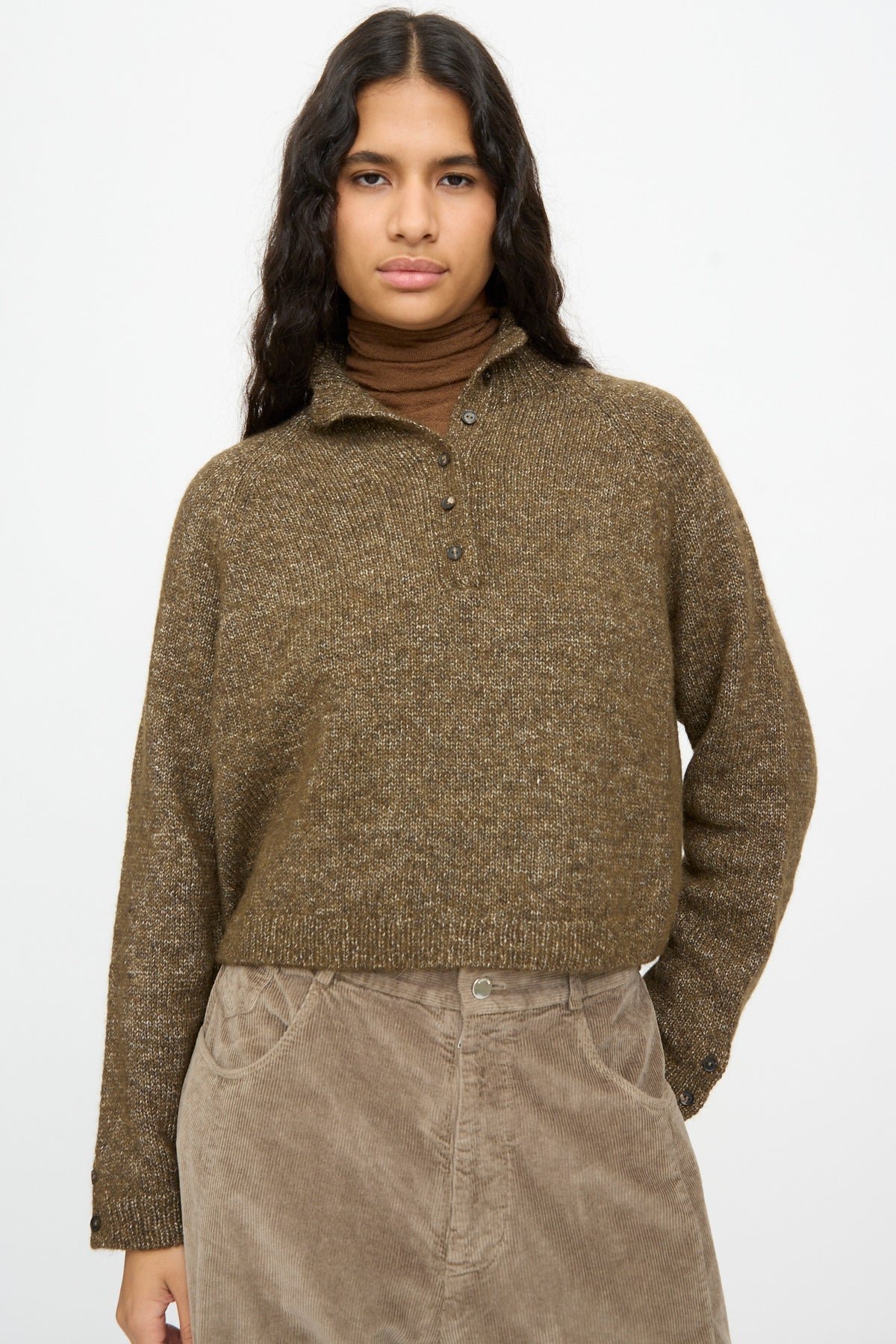 A person wearing a brown, speckled knit Melange Polo Sweater in Pardo over a brown turtleneck and light brown corduroy pants.