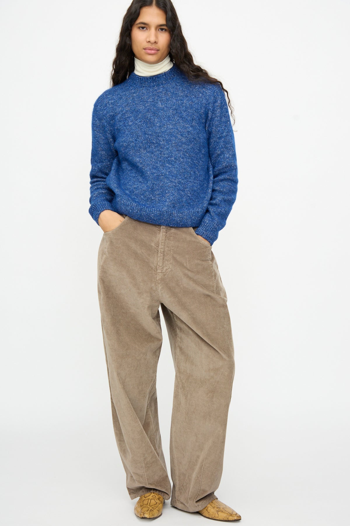 A person wearing a Cordera Melange Sweater in Blue over a white turtleneck, paired with wide-leg tan trousers and snakeskin-print pointed shoes.