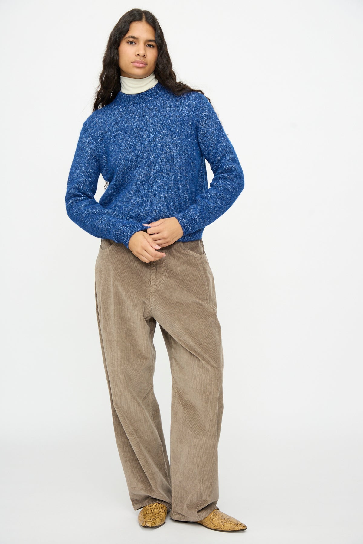 A person wearing a Cordera Melange Sweater in Blue over a white mock-neck top, paired with brown trousers and patterned pointed-toe shoes.