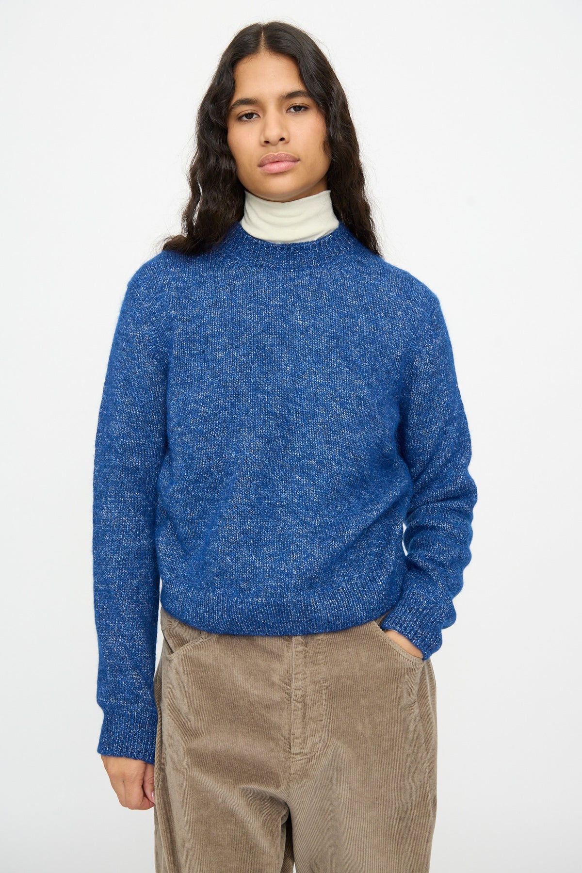 A model wears a relaxed fit Melange Sweater in Blue over a white turtleneck and tan corduroy pants.