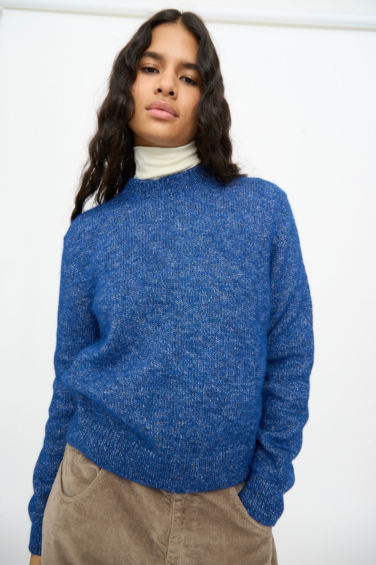 A person wears a relaxed fit Melange Sweater in Blue over a cream-colored turtleneck.