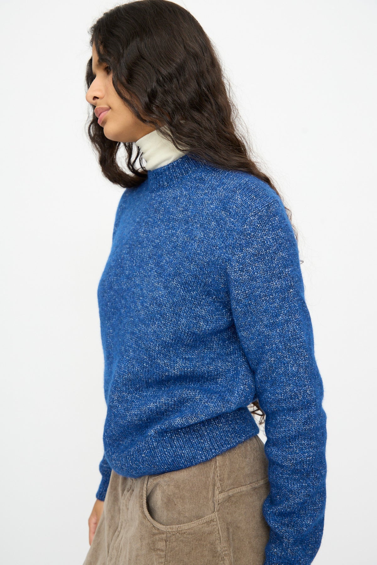 A person in profile wearing a Cordera Melange Sweater in Blue over a white turtleneck and tan trousers.